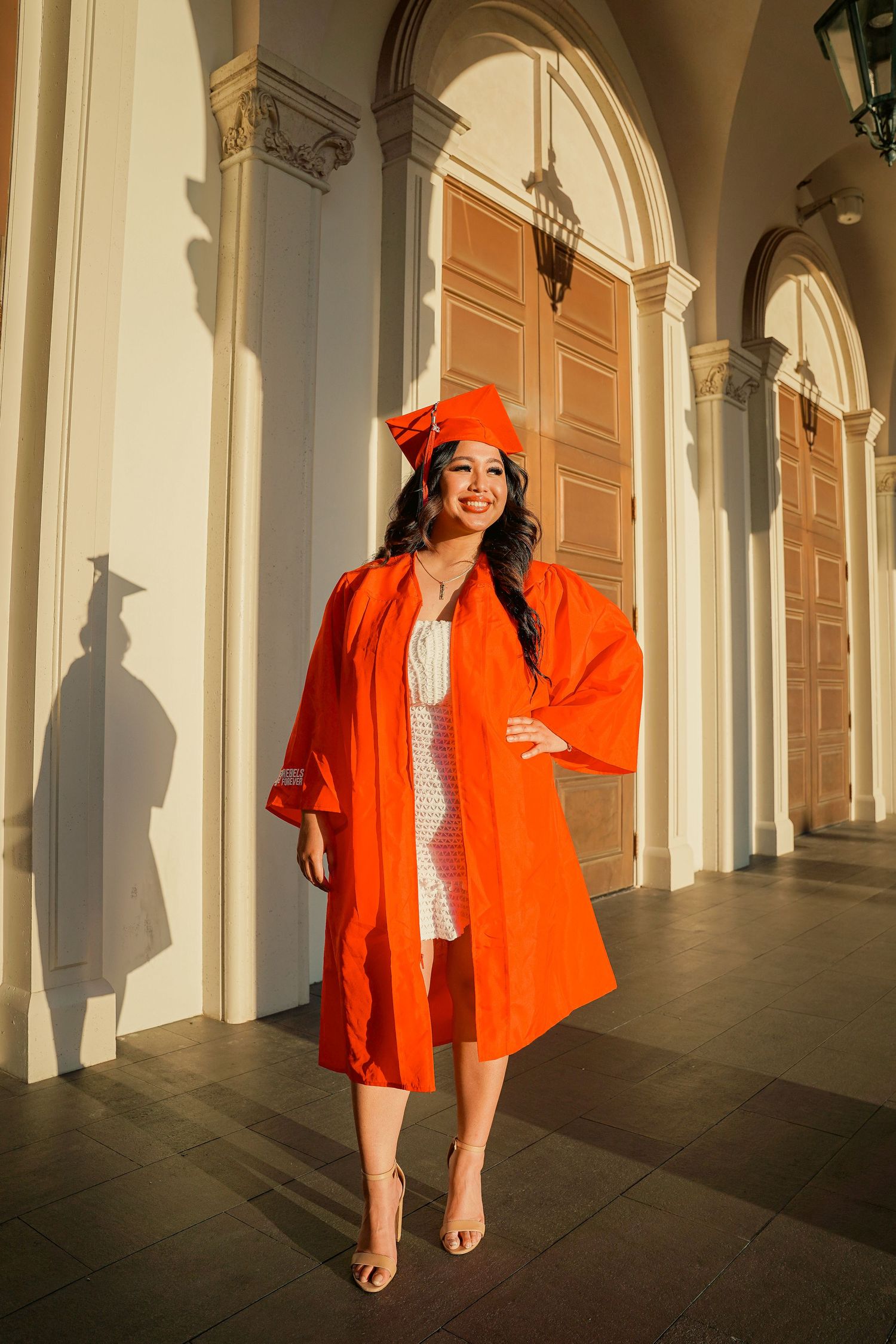 Graduate in orange cap and gown poses in sunlit colonnade with classical architecture.