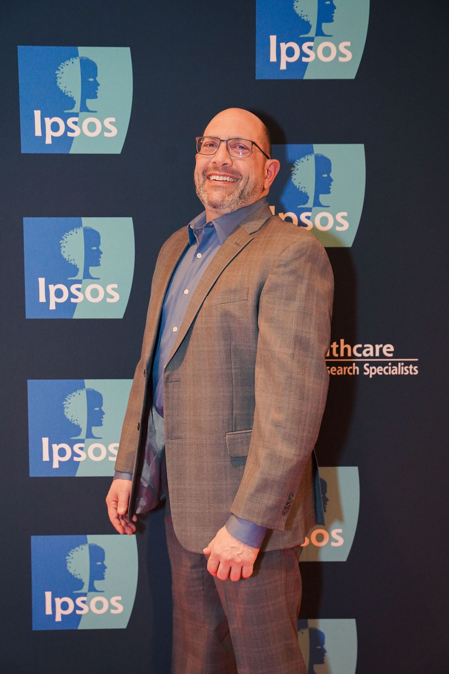 Smiling executive in suit jacket posing against Ipsos company logo backdrop.