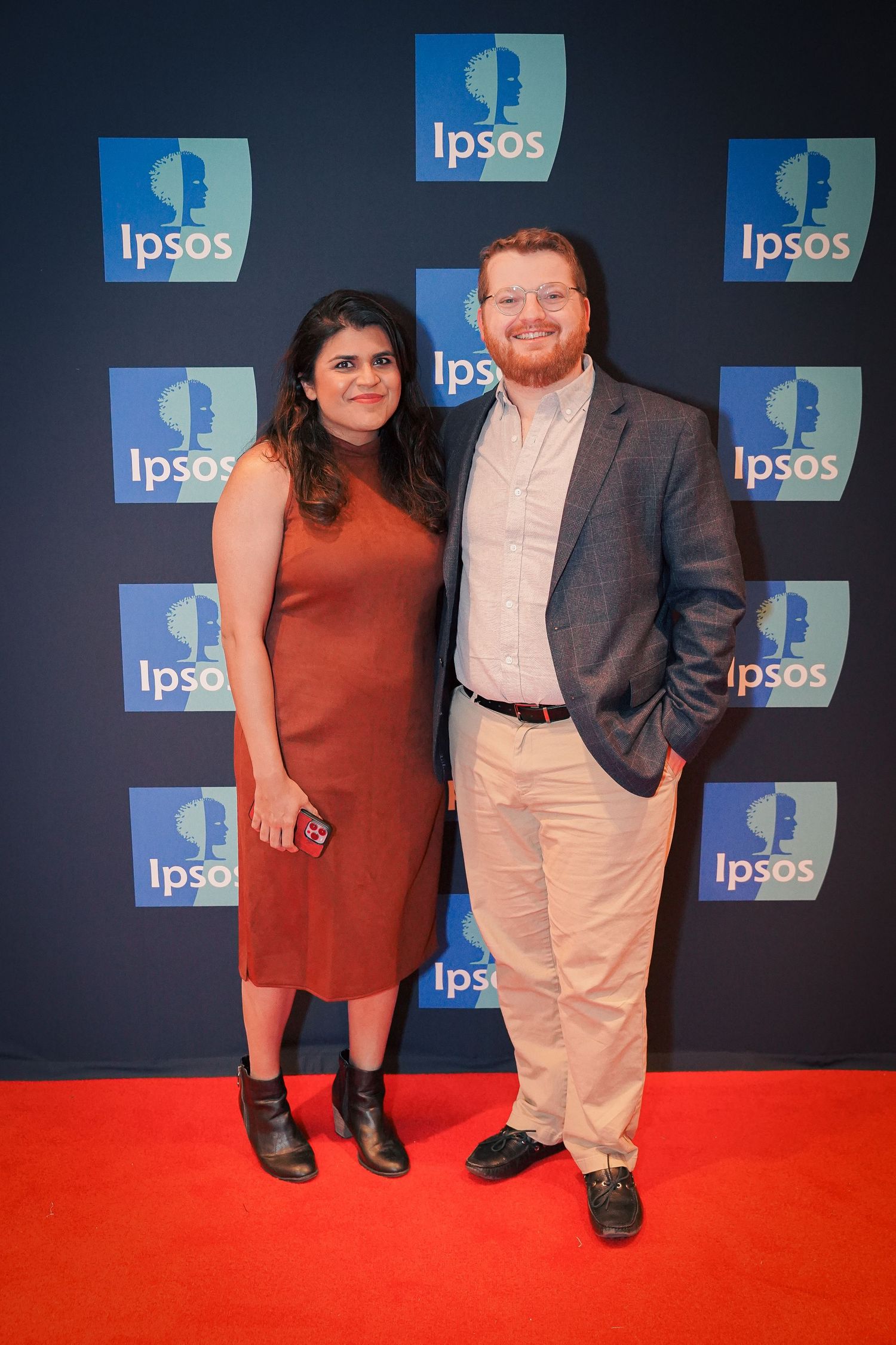A couple poses at an Ipsos event against a backdrop with red carpet and company logos.