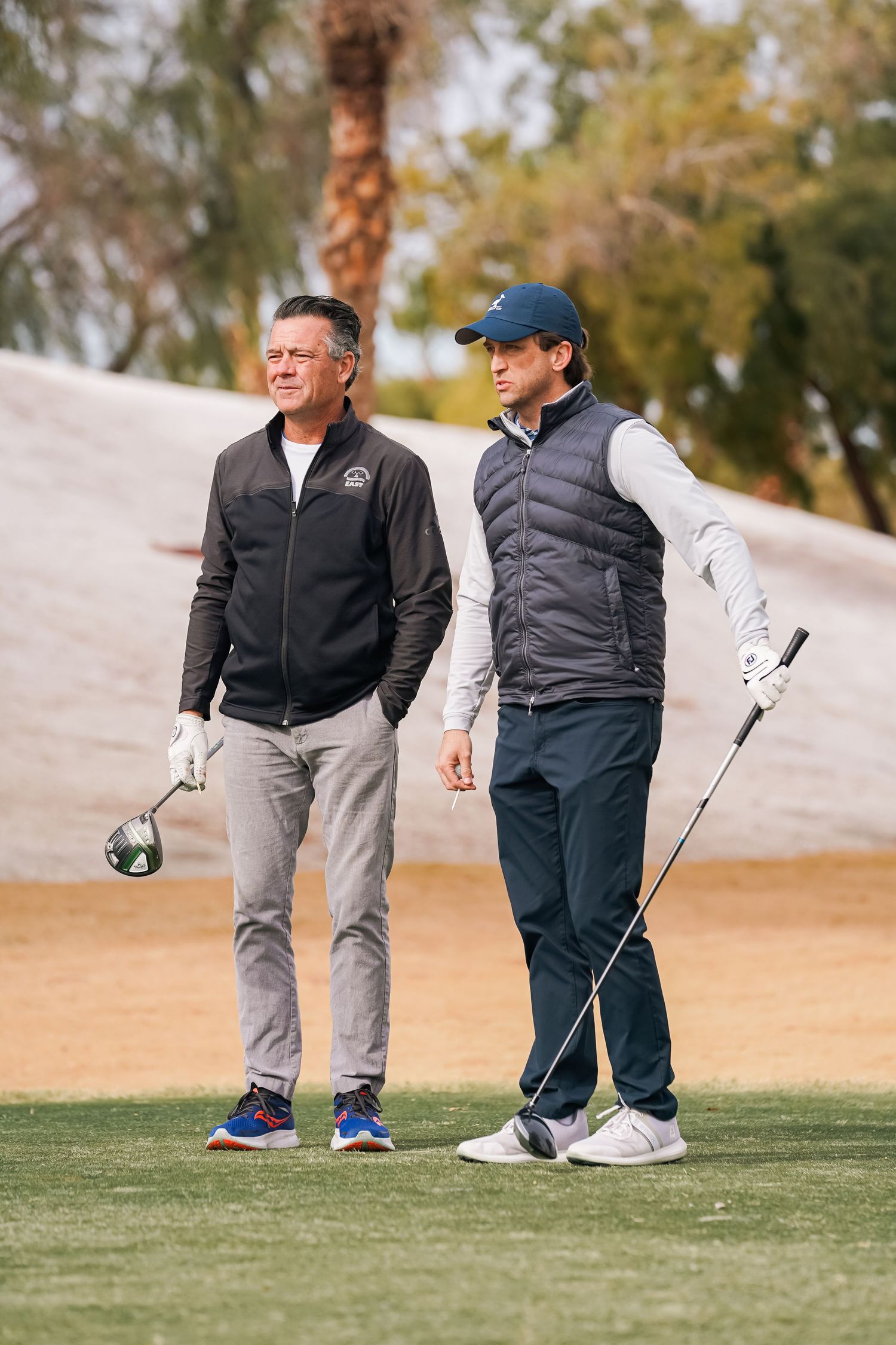 Two golfers in winter jackets standing with clubs on desert golf course.
