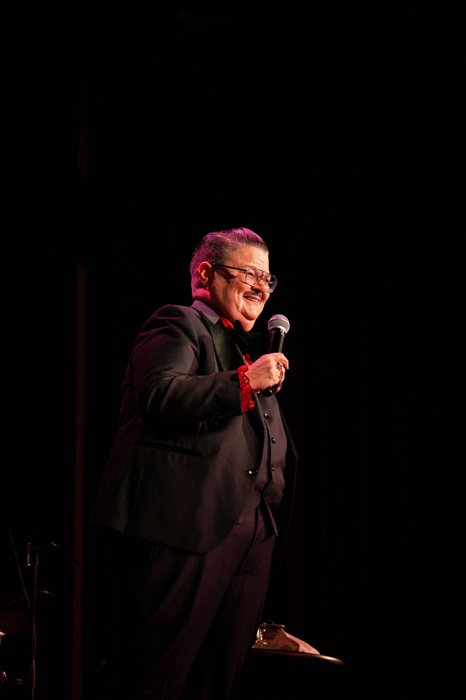 A performer in a dark suit speaks into a microphone on stage under dramatic lighting.