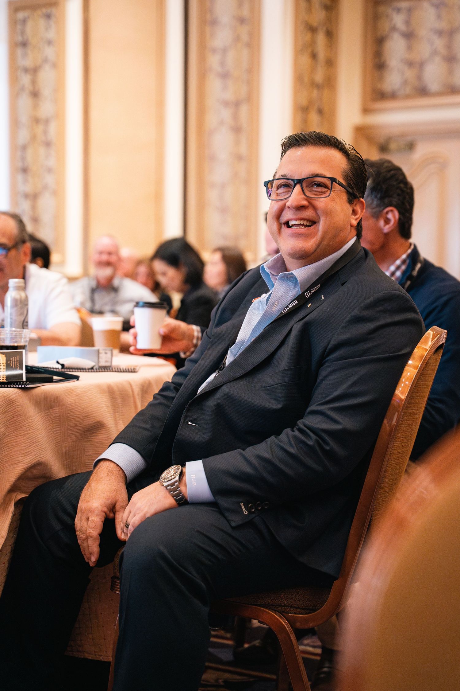 Conference attendee in suit and glasses smiling while seated at round table.