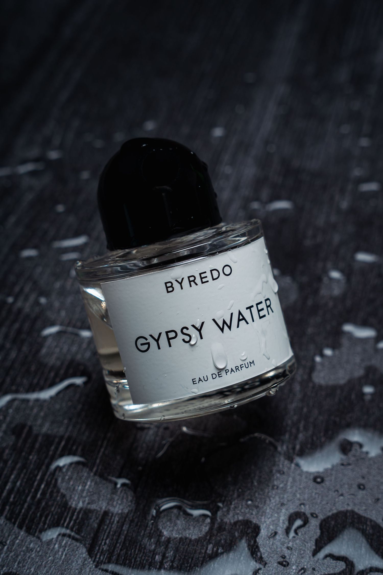 Byredo Gypsy Water perfume bottle on dark textured surface with water droplets.