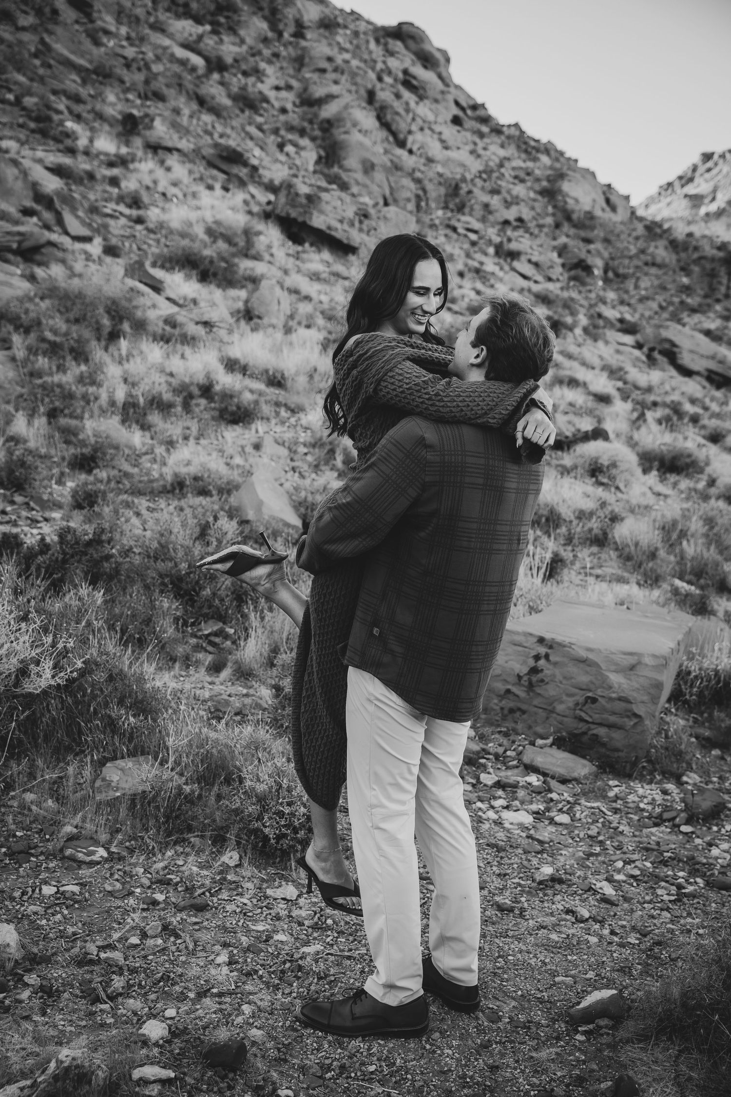 A joyful embrace between a couple against a rugged mountainous backdrop in black and white.