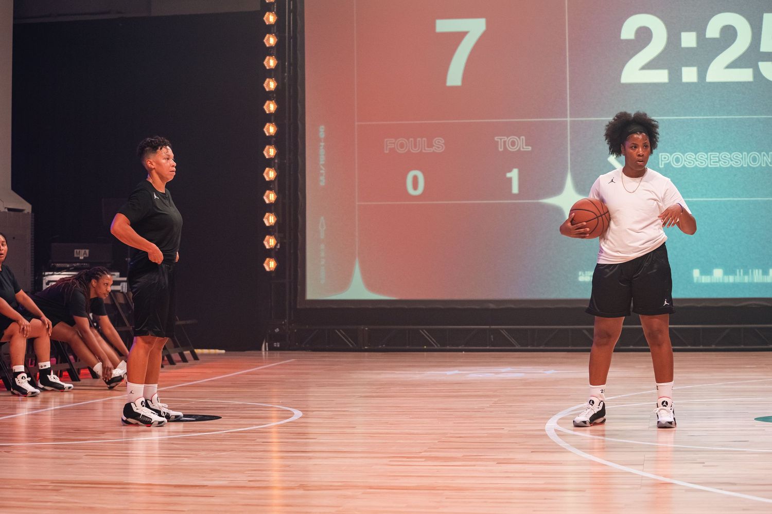 Basketball players practicing on indoor court with scoreboard showing game stats.