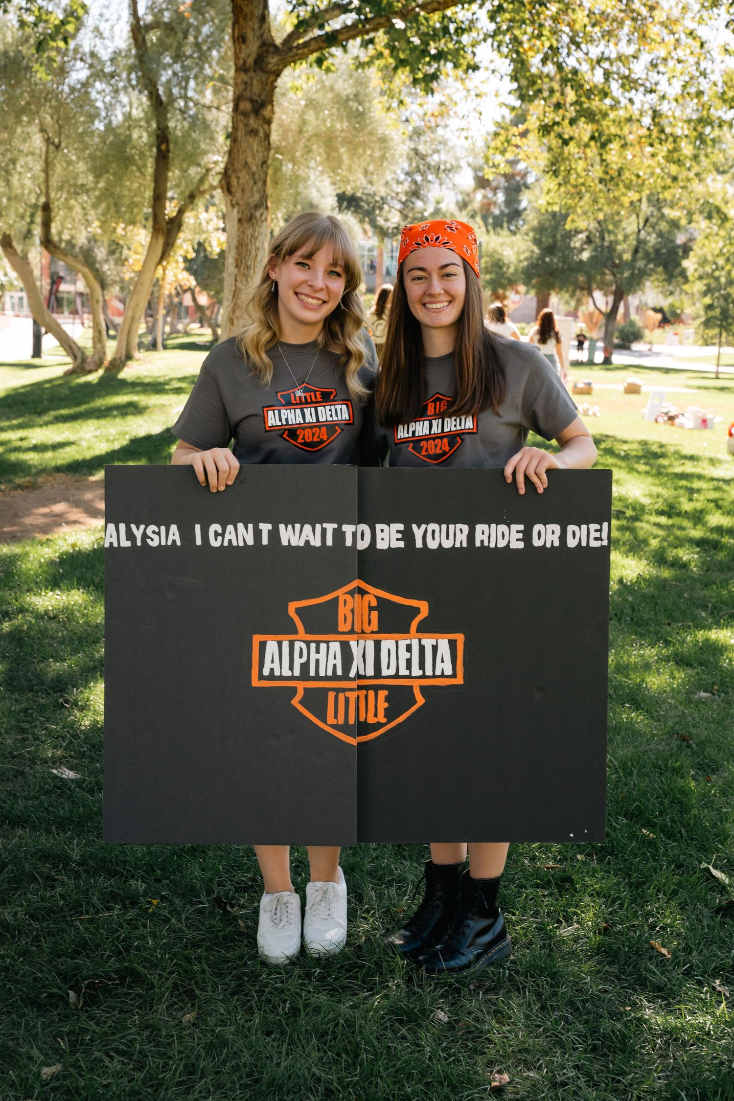 Two sorority members in matching dark t-shirts hold Alpha Xi Delta themed sign outdoors.