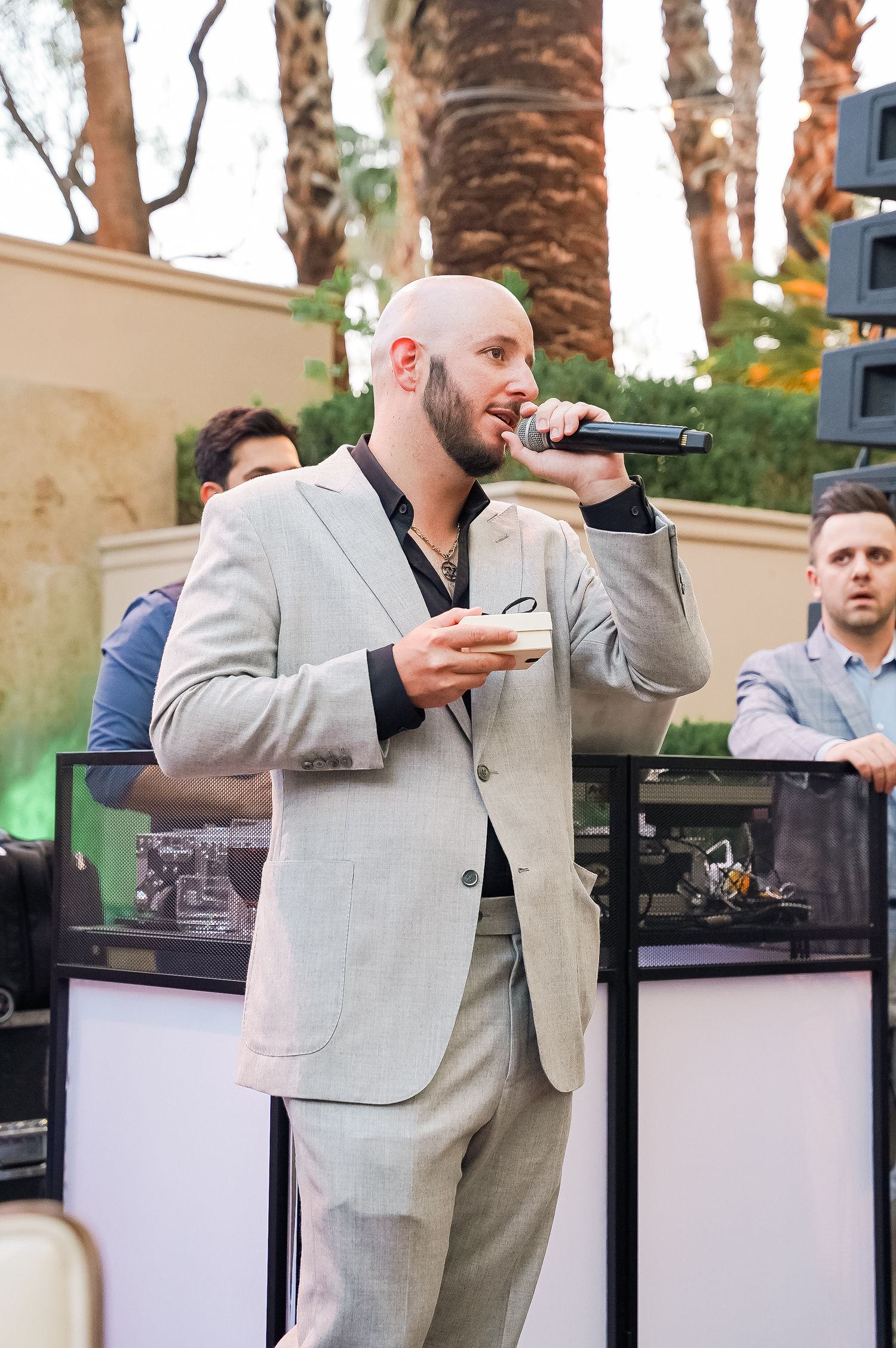 A performer in a light gray suit holds a microphone while speaking at an outdoor event with palm trees in the background.