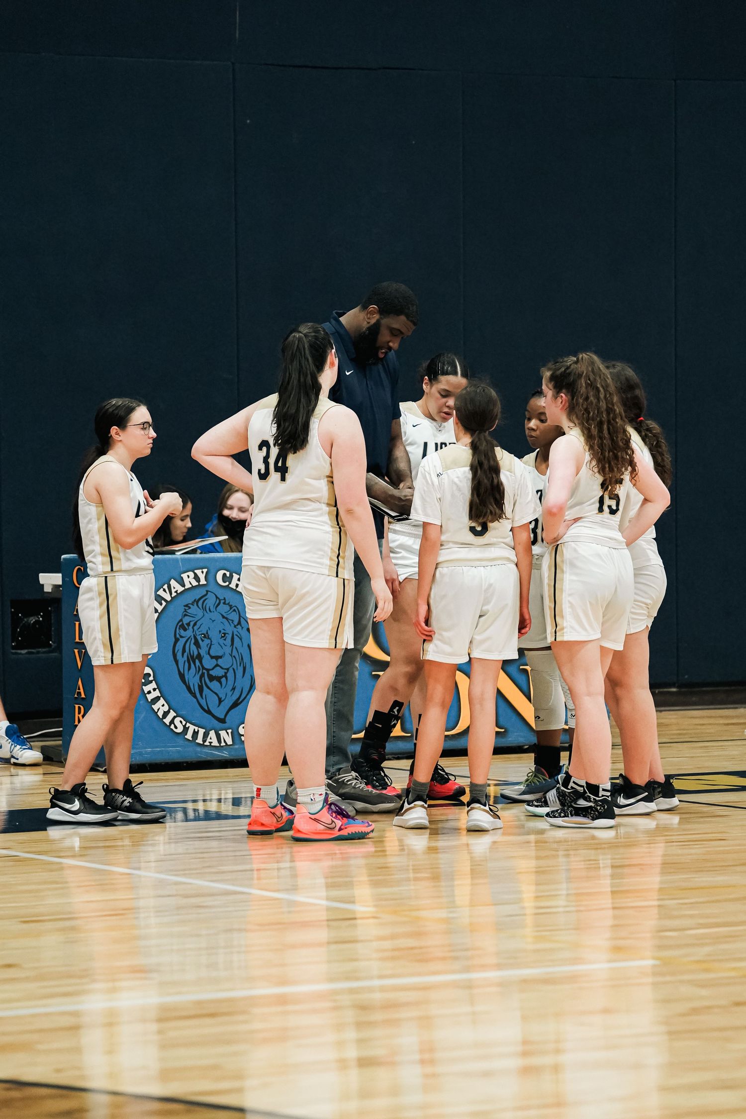 Basketball team huddles on court during timeout with school logo visible.