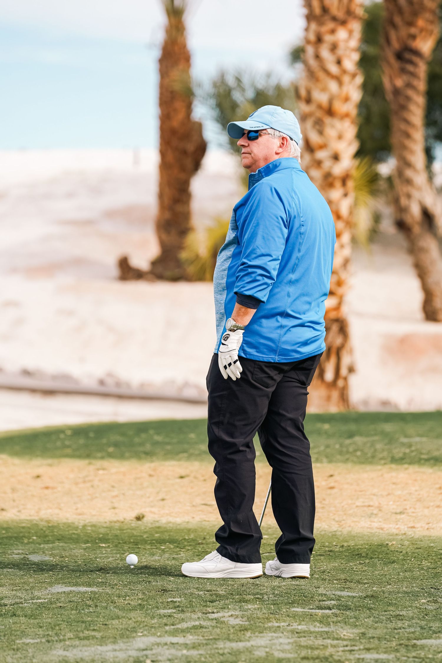 Golfer in light blue jacket stands on desert course next to golf ball.