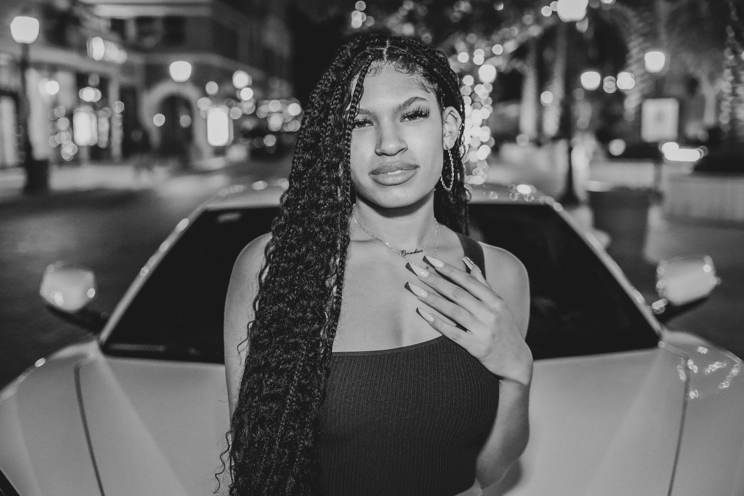 Black and white portrait of woman with long braids posing in front of car at night.