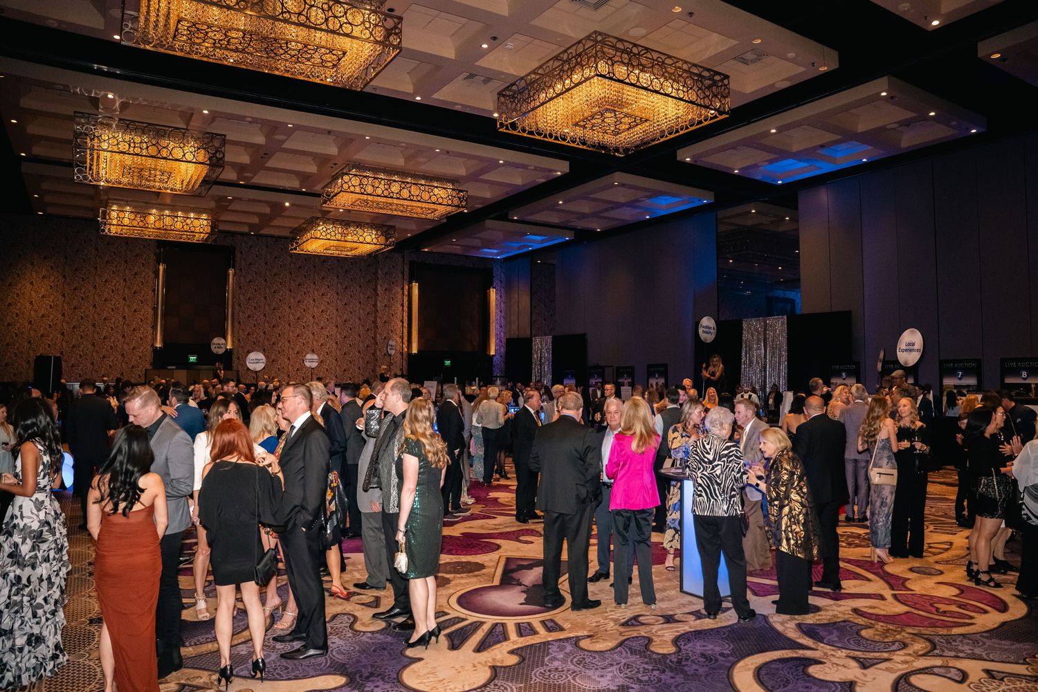 Large crowd of formally dressed attendees mingling at an elegant ballroom event with crystal chandeliers.