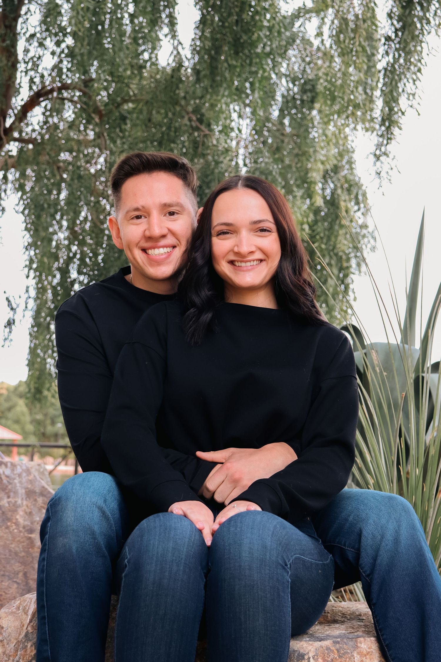 Happy couple in matching black sweaters embrace while sitting outdoors near willow trees.