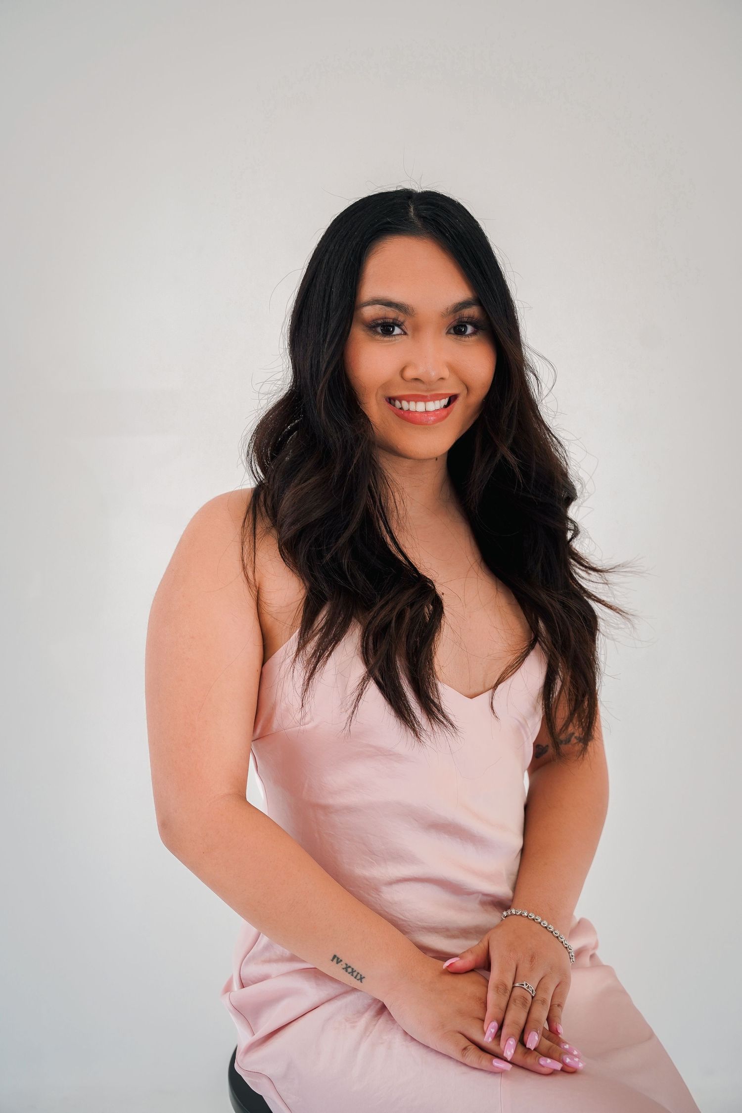 Woman in pink slip dress seated on black chair against white background.