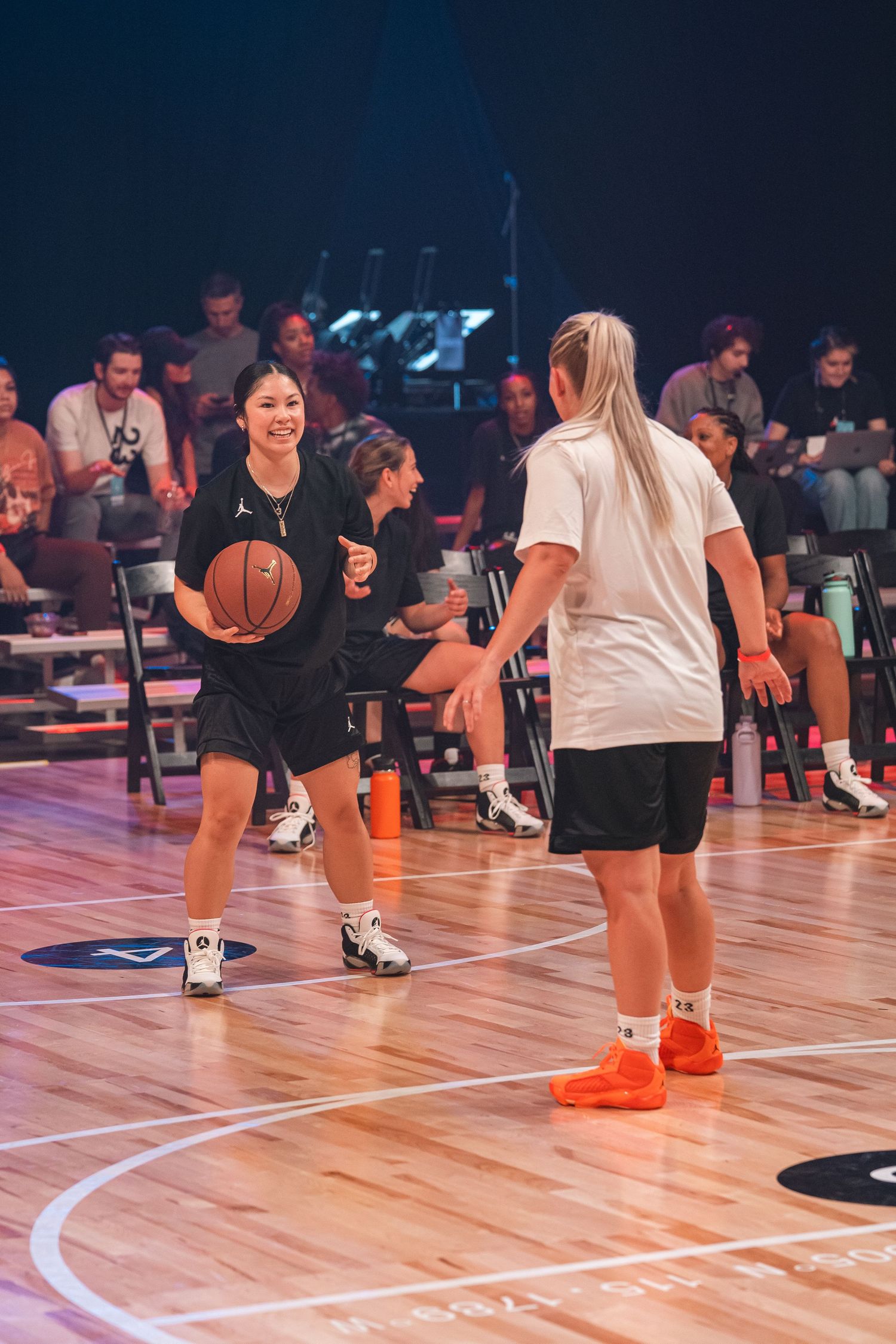 Two players practicing basketball drills on indoor court with spectators in background.