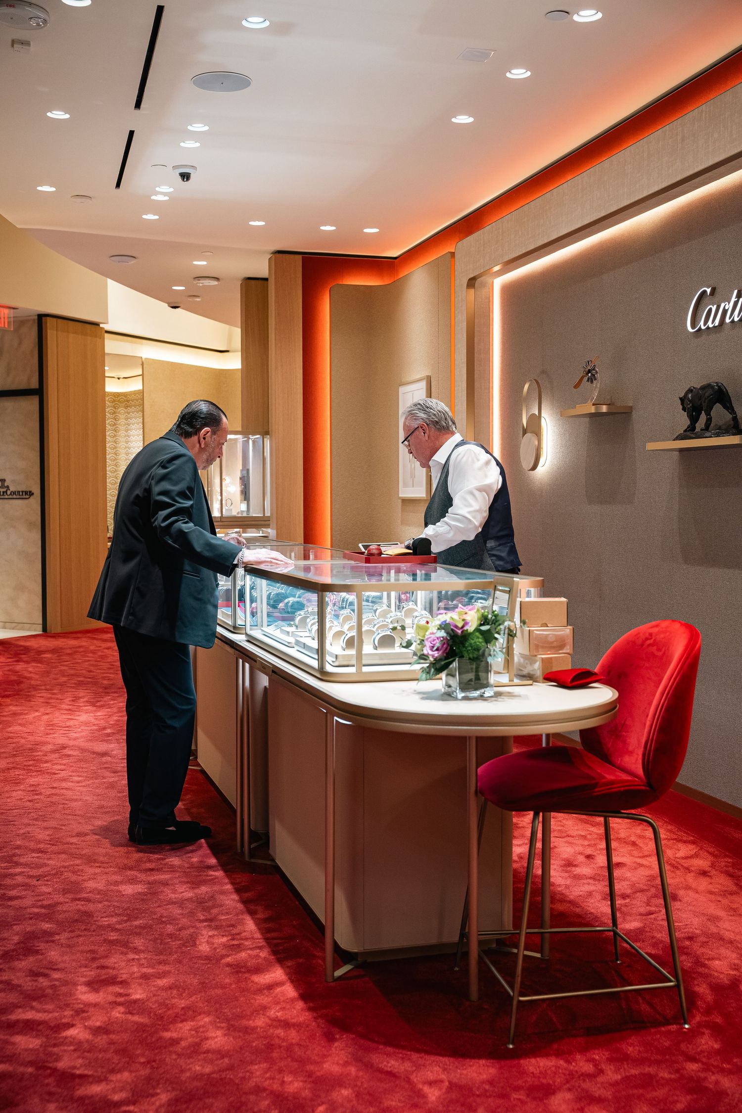 Customers examine jewelry displays in a luxury retail store with bright red carpet and modern lighting.