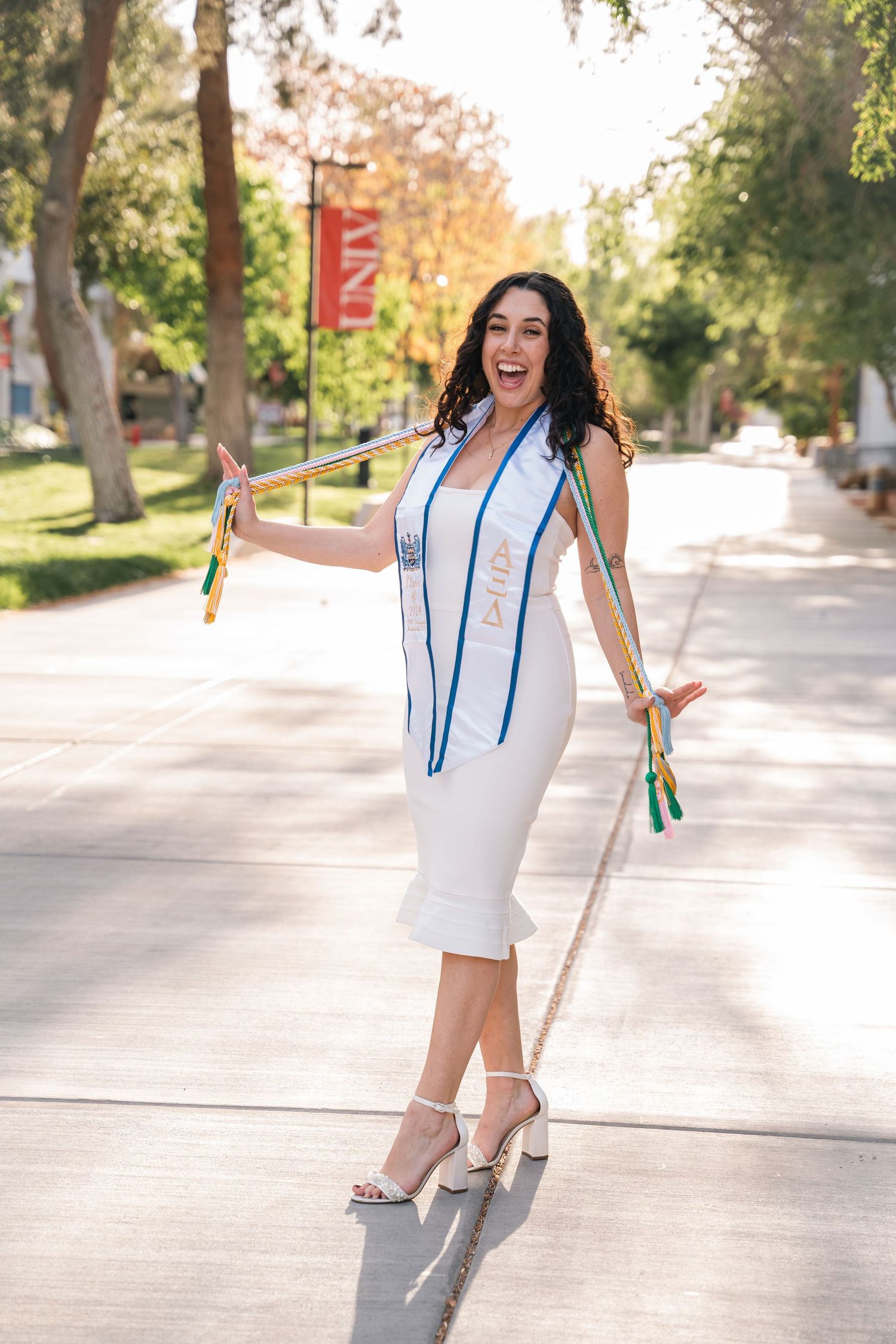 A graduate in a white dress and graduation sash poses joyfully on a sunny campus walkway.