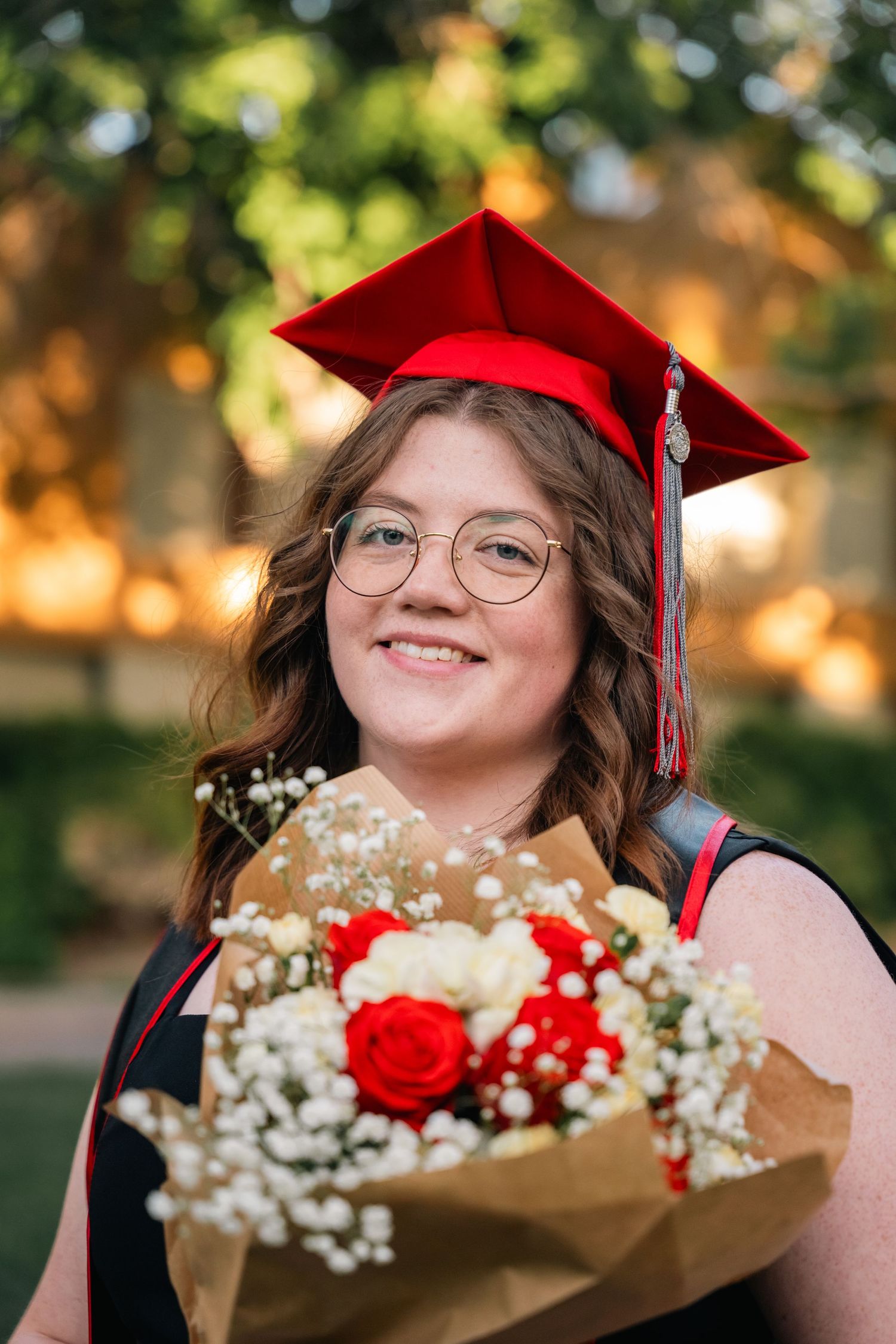 Graduate in red cap holding bouquet of roses and baby's breath flowers smiles at camera.