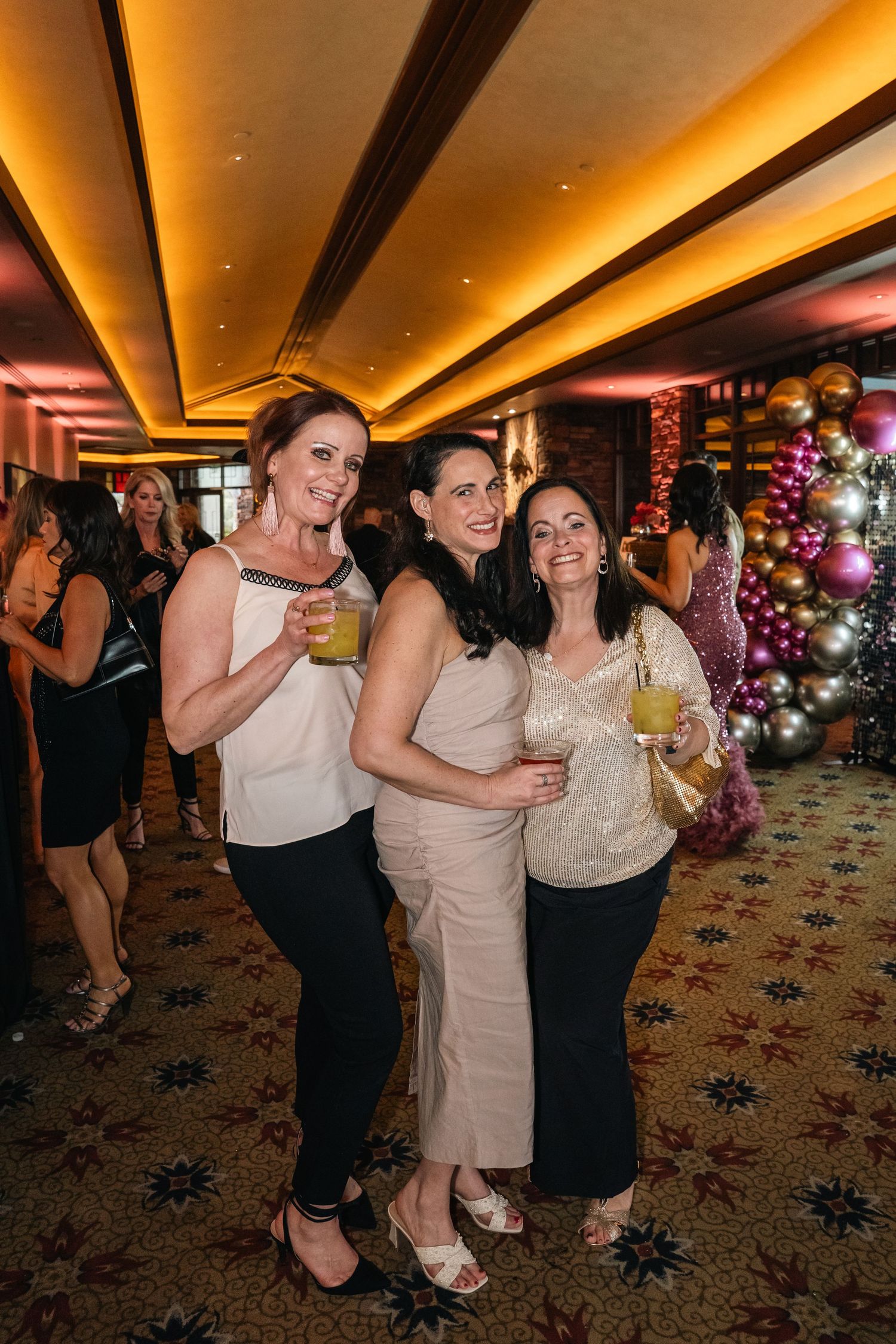 Three friends in evening wear pose with drinks at a party with warm lighting.