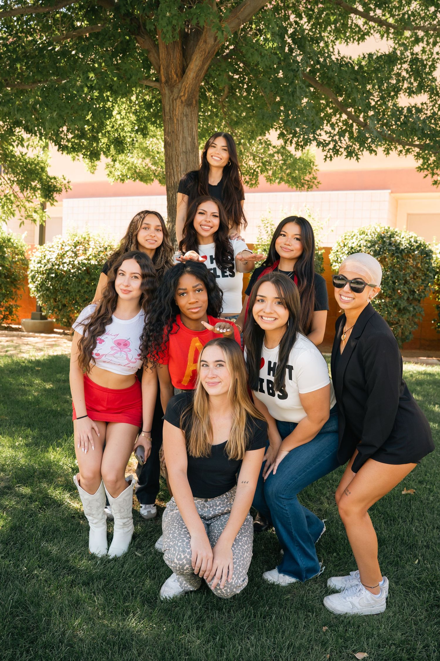 Group of friends in casual attire pose together on campus lawn during a sunny day.
