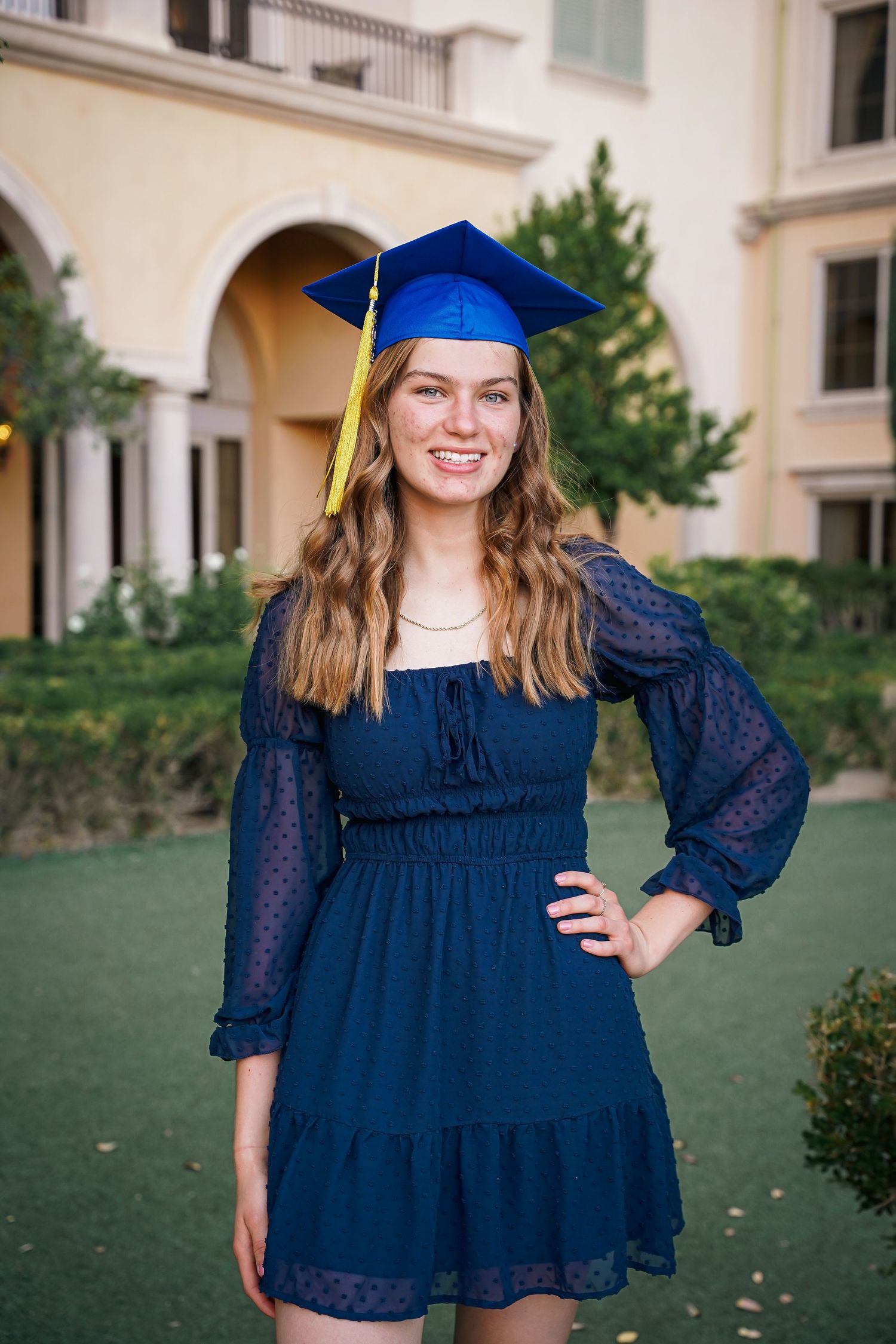 Graduate in navy blue dress and cap poses on campus lawn.