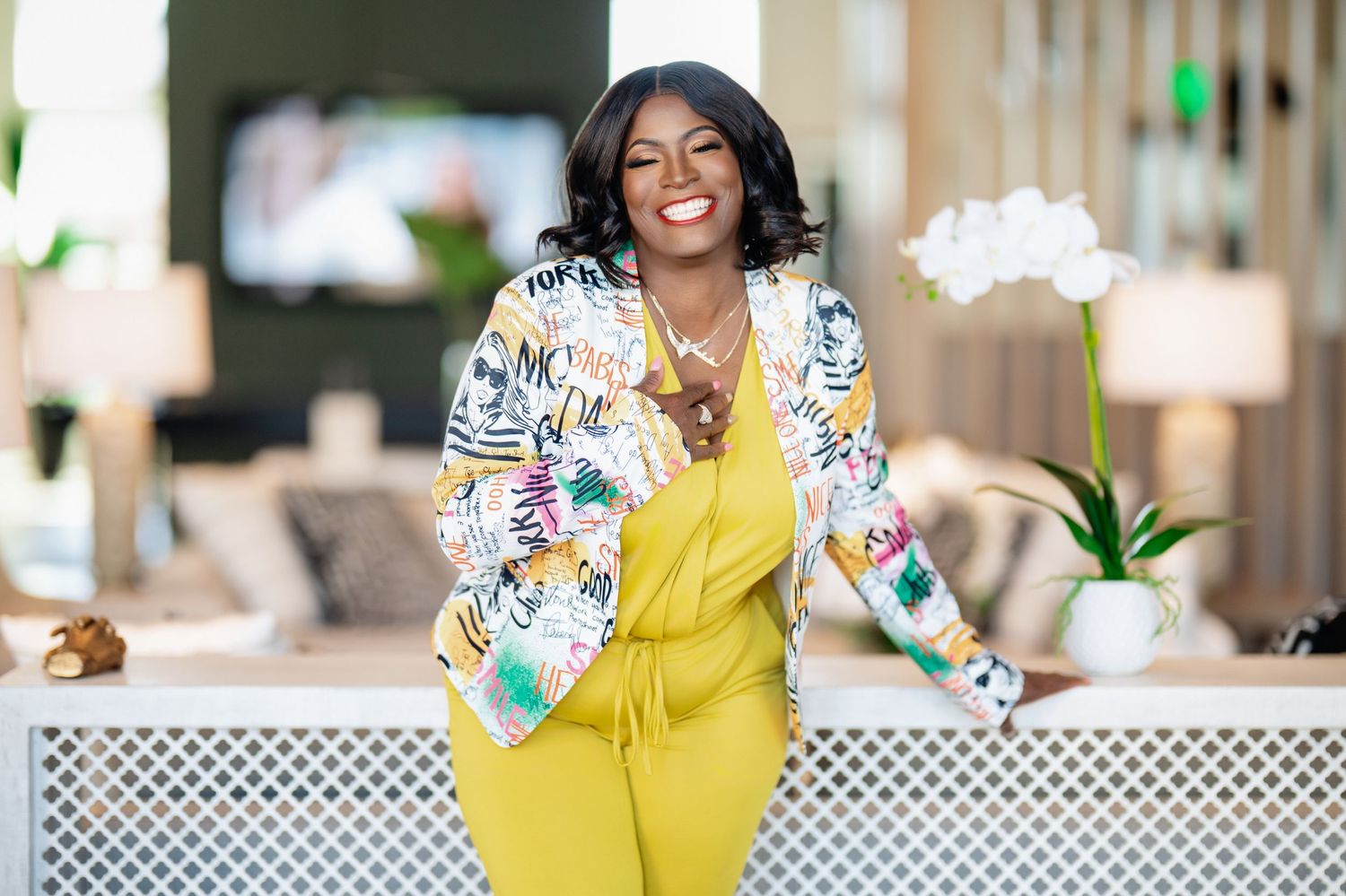 Smiling woman in yellow jumpsuit and floral jacket posing confidently in a bright, modern interior setting.