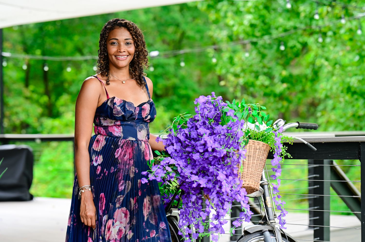 Woman in floral dress smiling beside a bicycle adorned with purple flowers and basket outdoors.