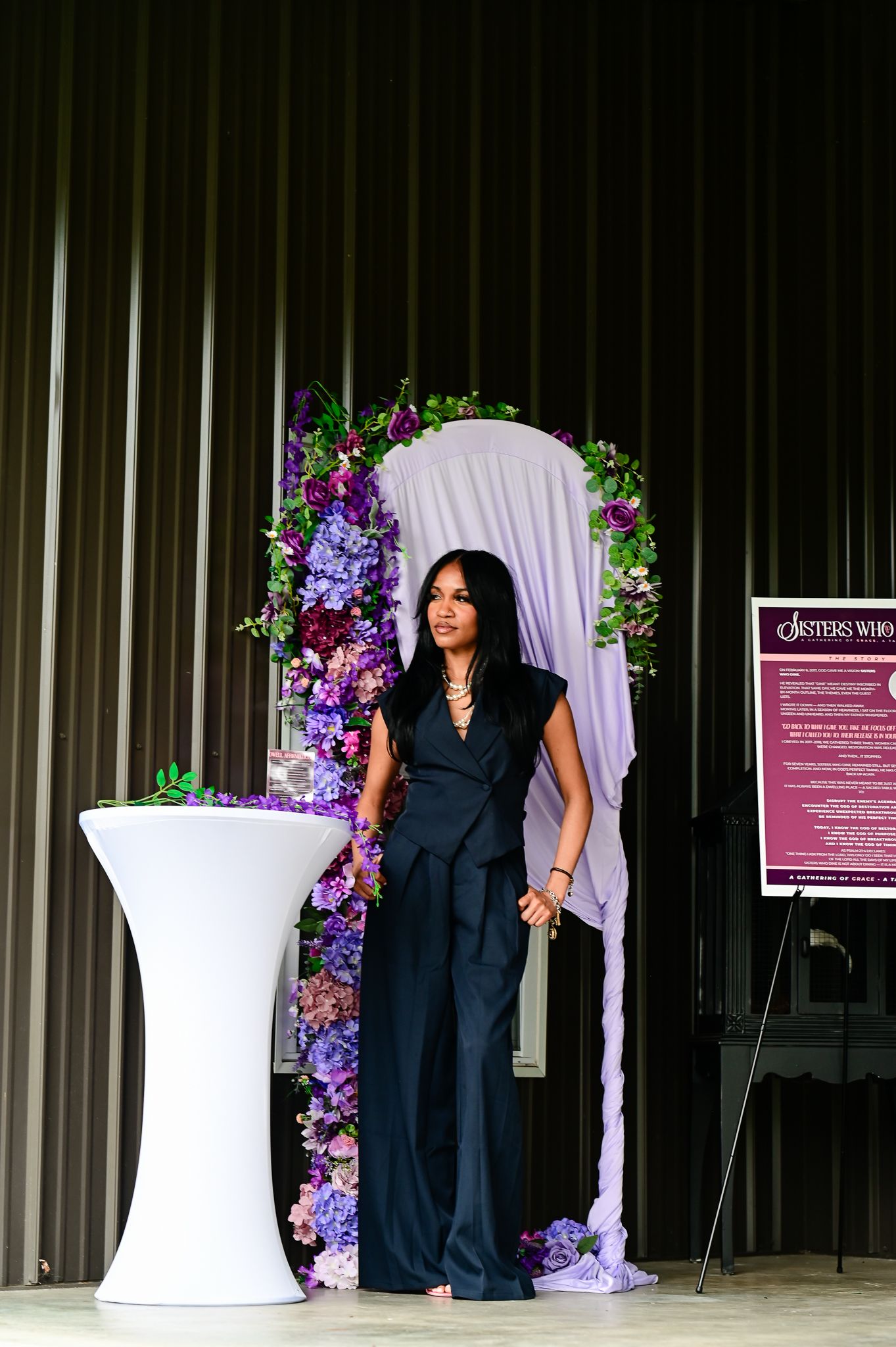 Woman in black jumpsuit stands before a floral arch with purple flowers at an elegant event.