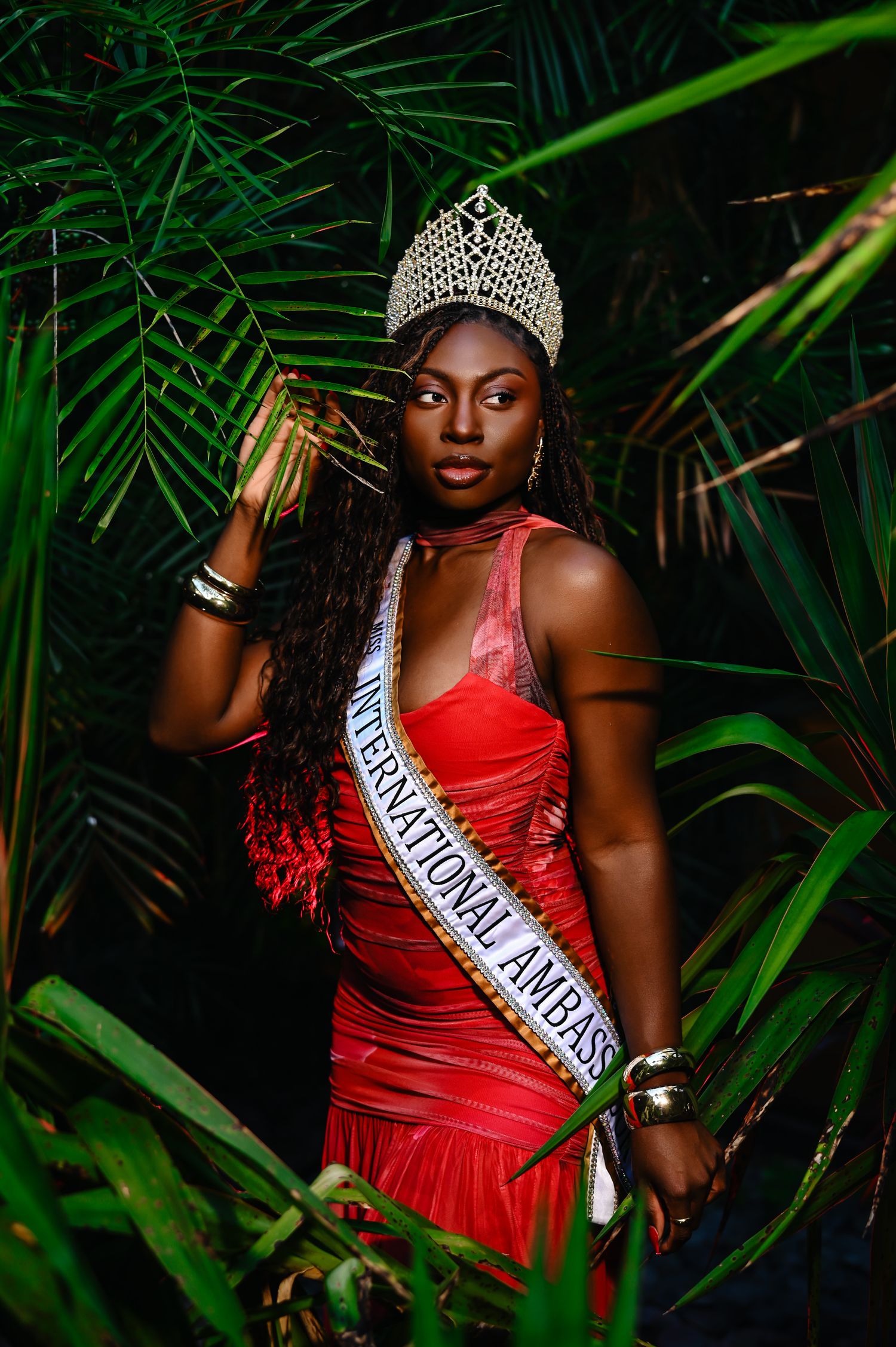 A pageant contestant in a red evening gown poses elegantly against a backdrop of tropical palm leaves.
