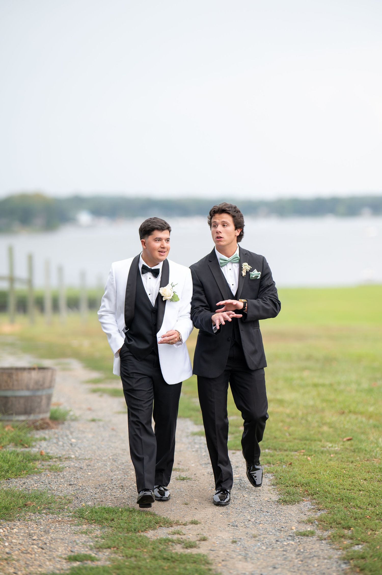 Two groomsmen in formal attire walk along a waterfront path during a wedding celebration on a cloudy day.