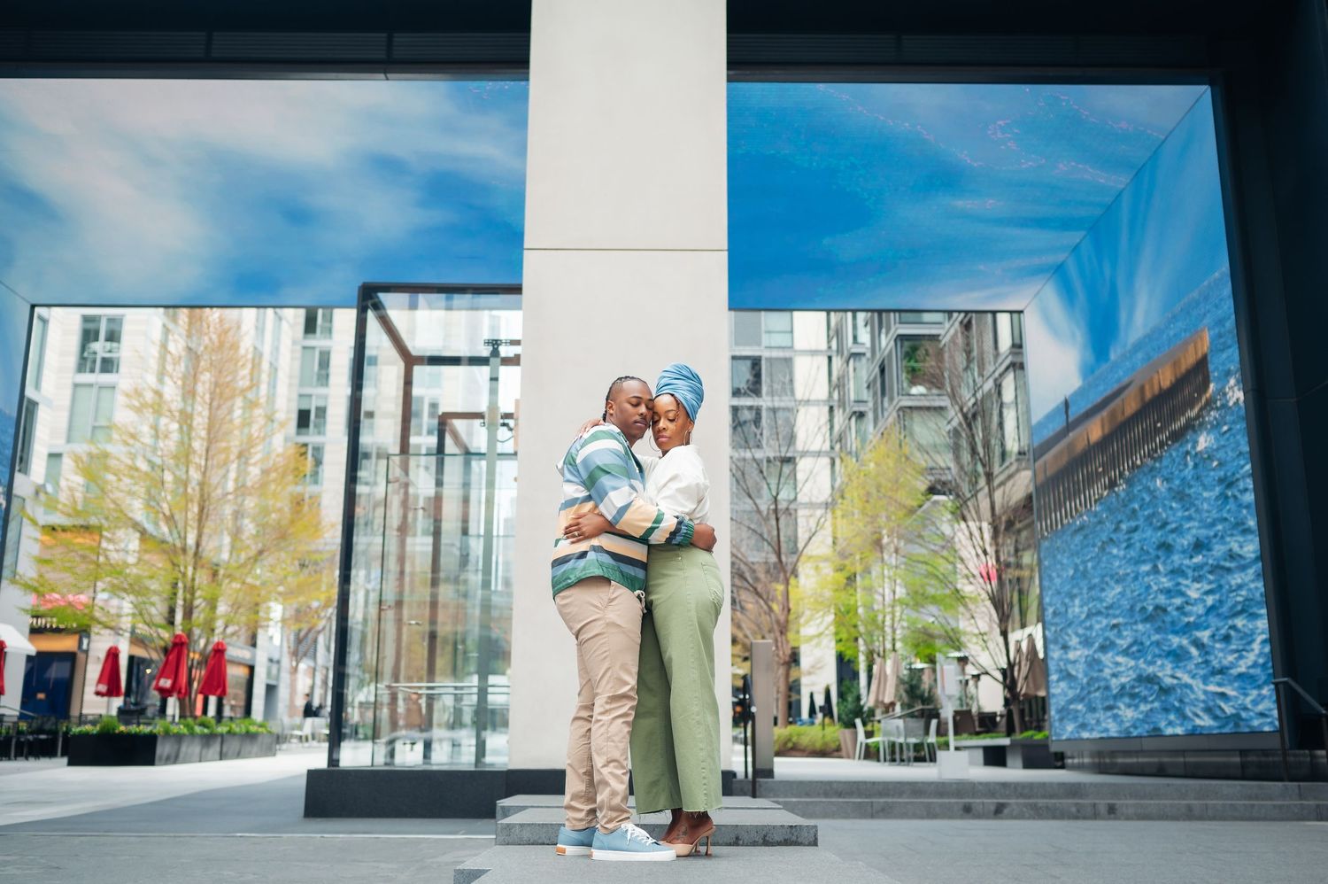 Two individuals embrace in a sweet moment outdoors near modern buildings with white columns and yellow autumn trees.