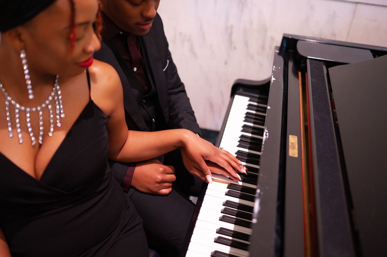 Two musicians perform together at a black grand piano, with one wearing an elegant gown and sparkling necklace.
