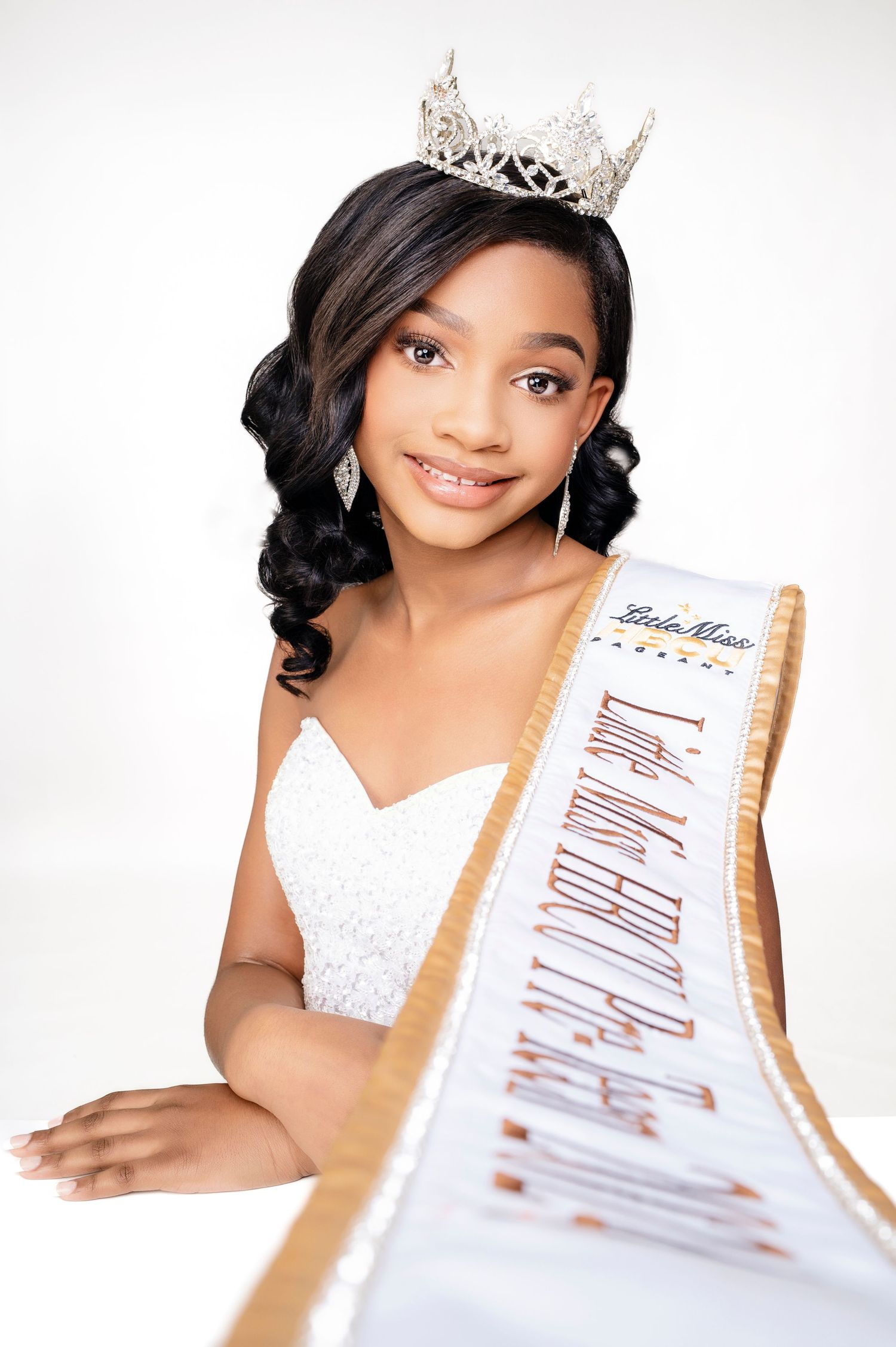 A beauty pageant contestant wearing a white gown, sparkling crown and sash poses for an elegant portrait.