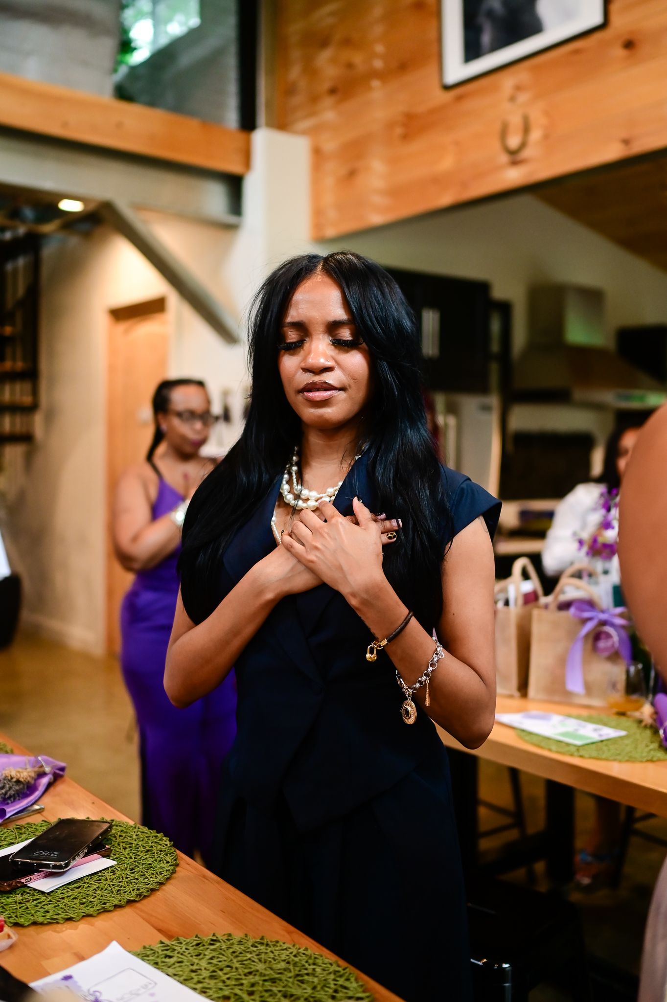 Woman in black dress touching necklace at indoor gathering with guests in purple attire in background.