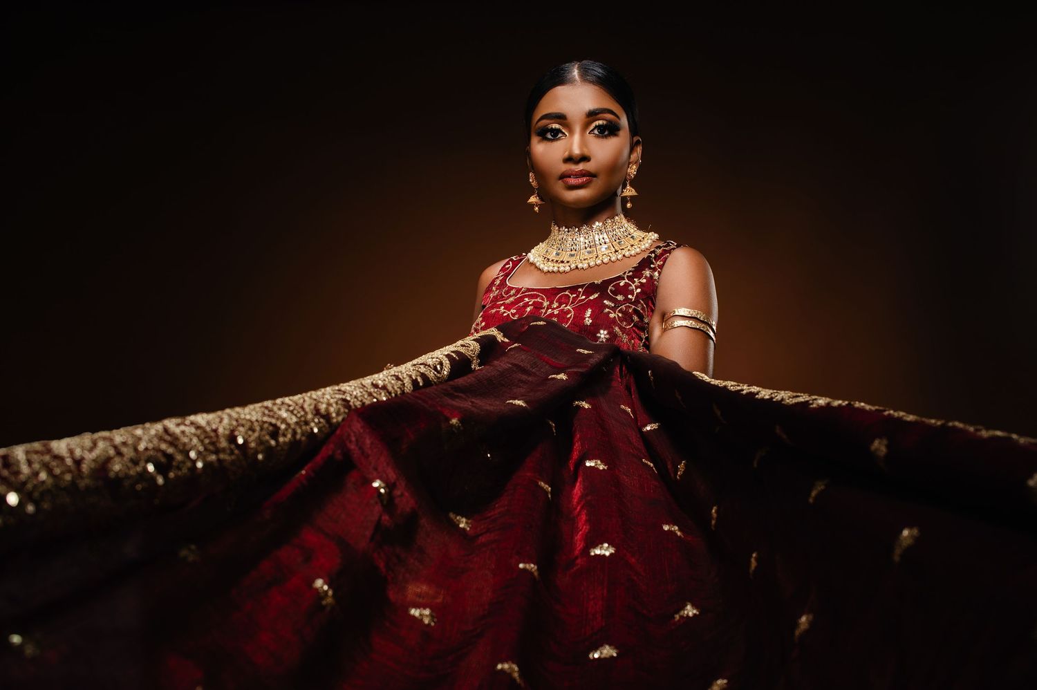 A woman in a stunning crimson bridal sauri with gold embroidery poses gracefully against a dark background.