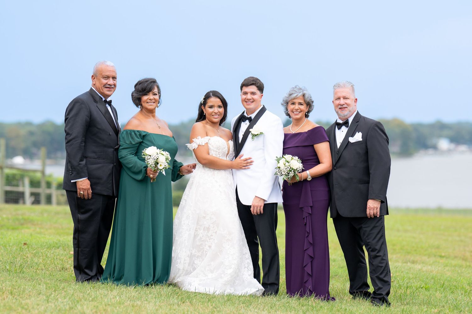 A wedding party poses on a grassy hillside overlooking water on a bright sunny day.