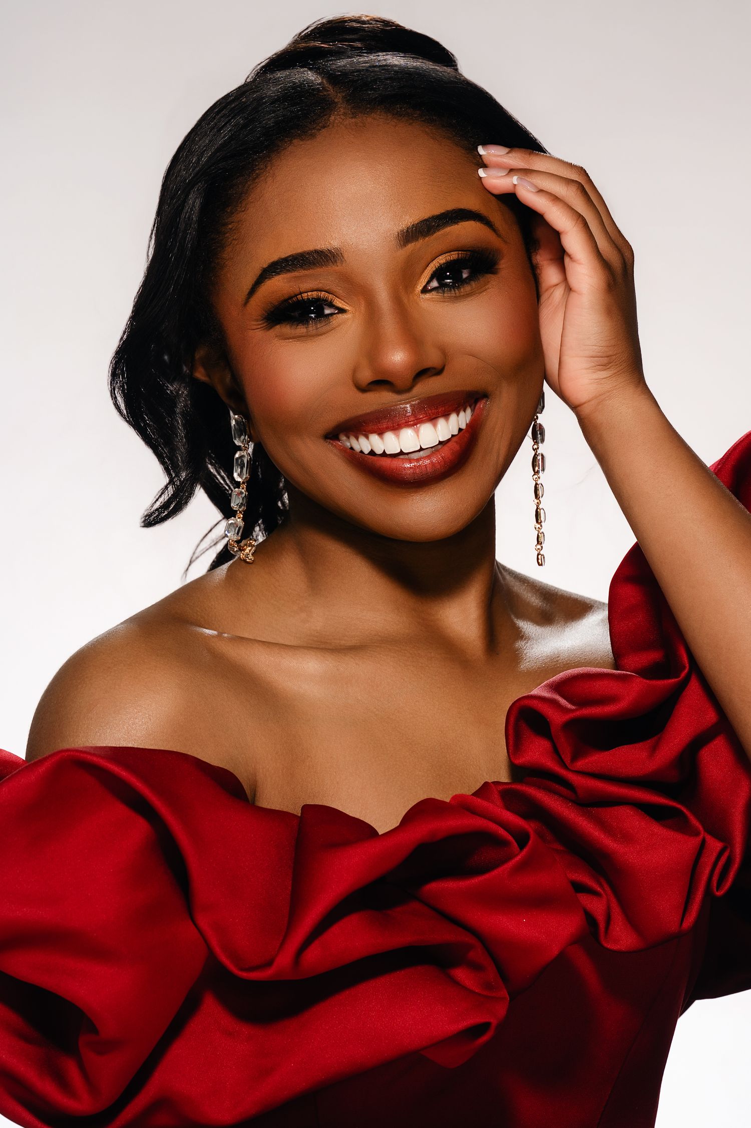 Smiling woman in red off-shoulder ruffled gown with elegant drop earrings poses confidently against white background.