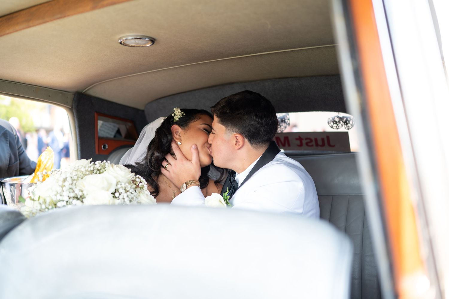 A romantic moment between newlyweds sharing a kiss in the back of a classic car decorated with flowers.