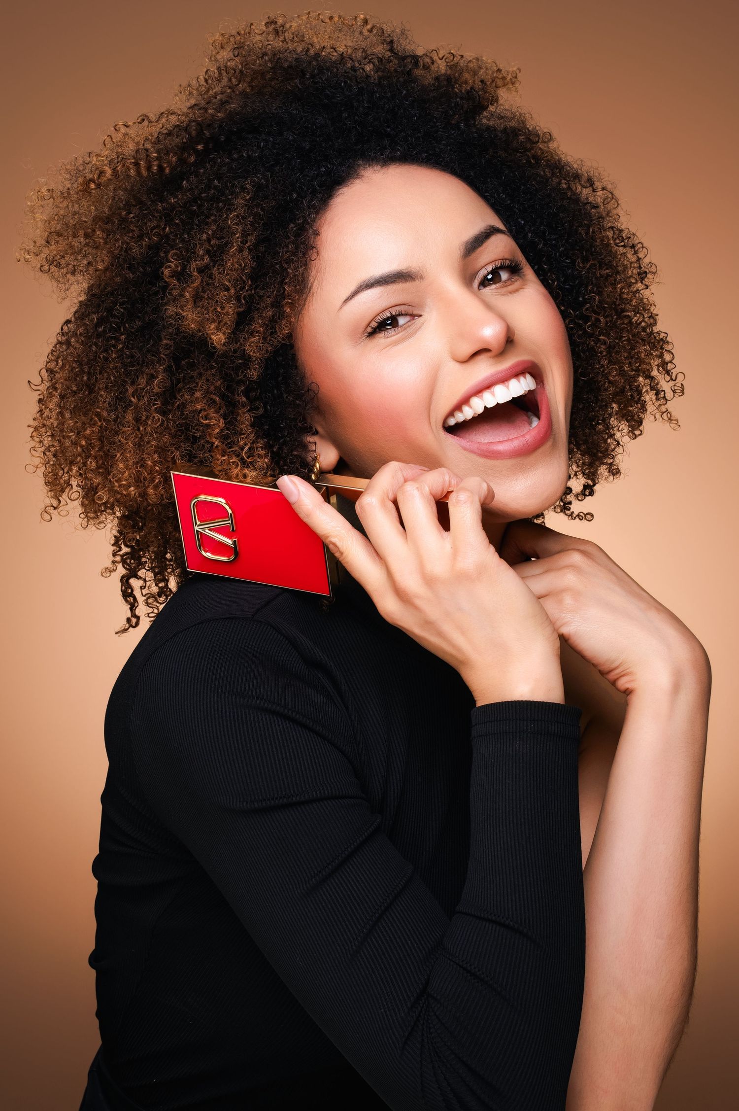 Woman with natural curly hair wearing black sweater laughs while holding red McDonald's card against brown background.