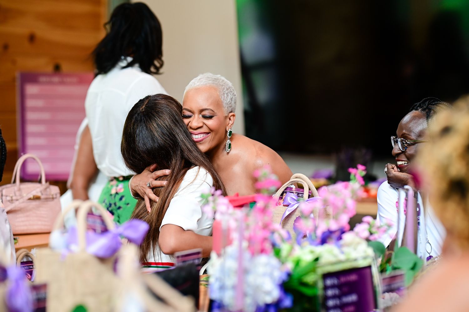 Two women sharing a warm hug at a decorated event table with colorful floral arrangements.