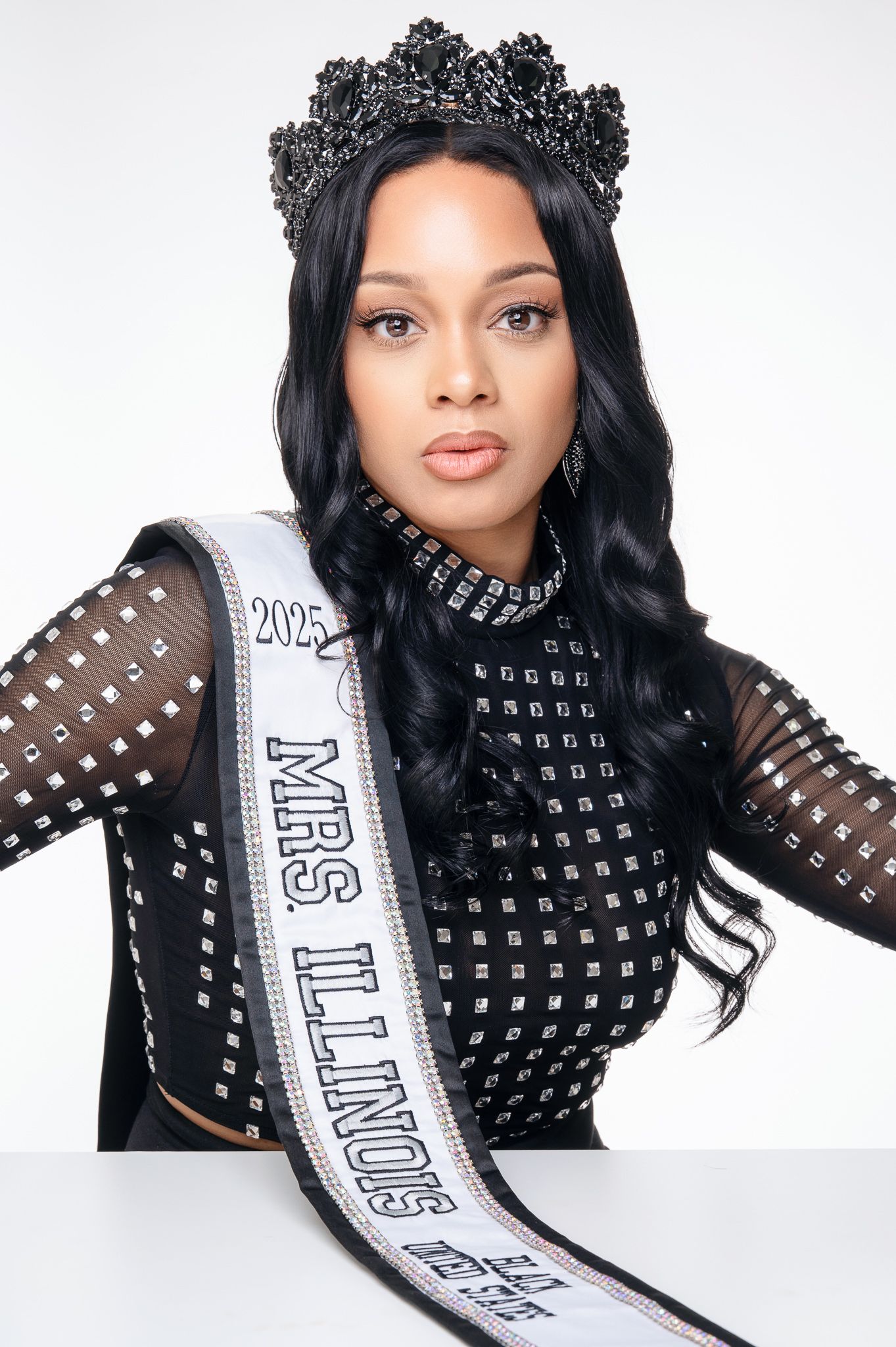 A pageant contestant wearing a black studded top, ornate crown and Miss Illinois sash poses for a formal portrait.