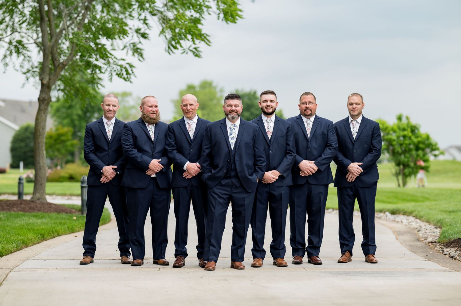 A group of groomsmen in matching navy suits standing on a path outdoors.