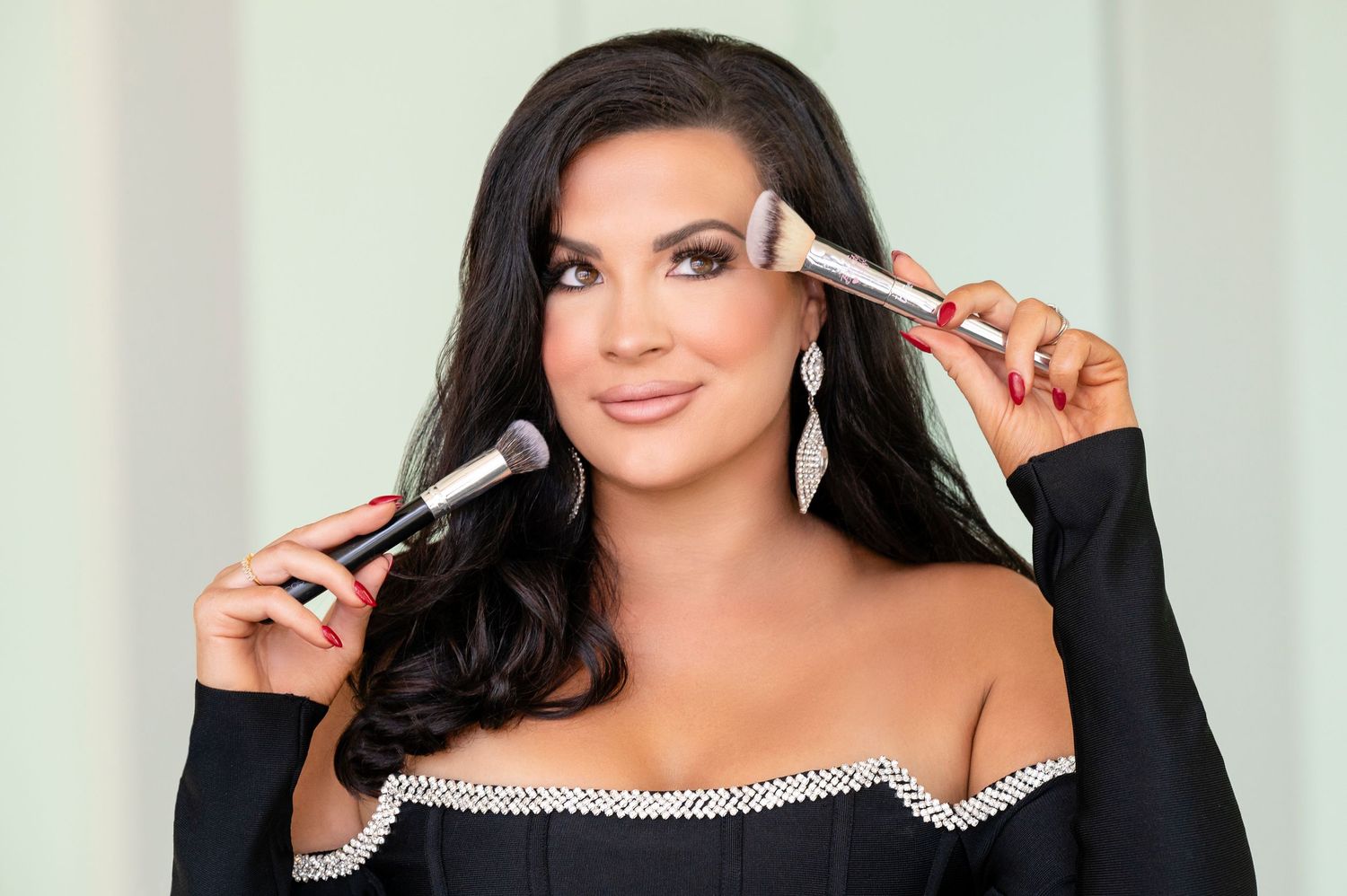 A beauty professional holds makeup brushes while wearing a black rhinestone-trimmed dress.
