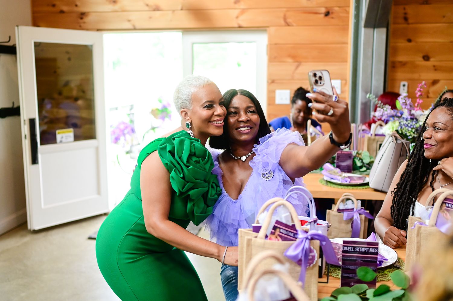 Two women take a selfie at a purple-themed party in a decorated venue with gifts on the table.