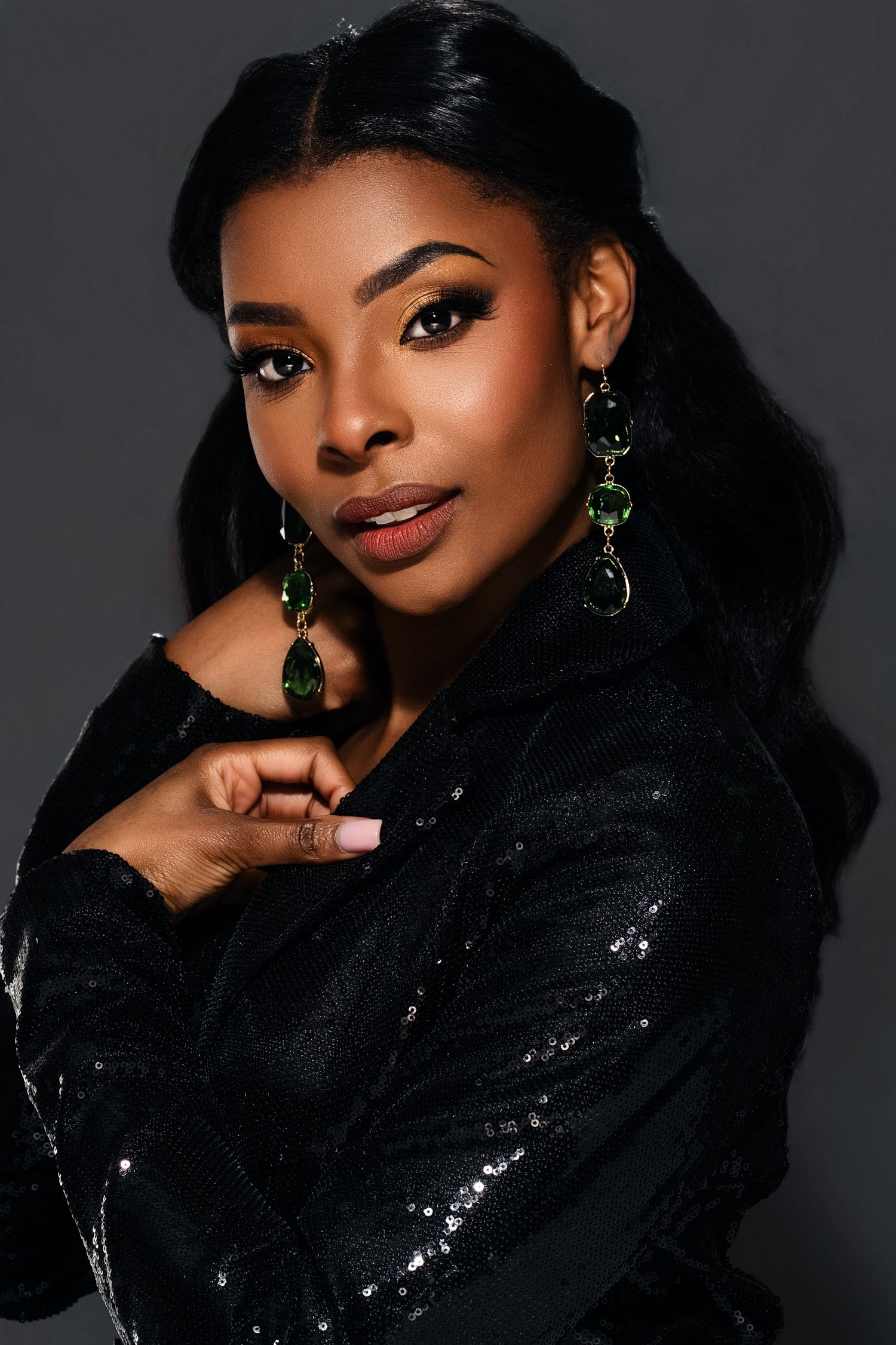 Elegant woman in black sequin jacket with green gemstone earrings poses against dark gray background.