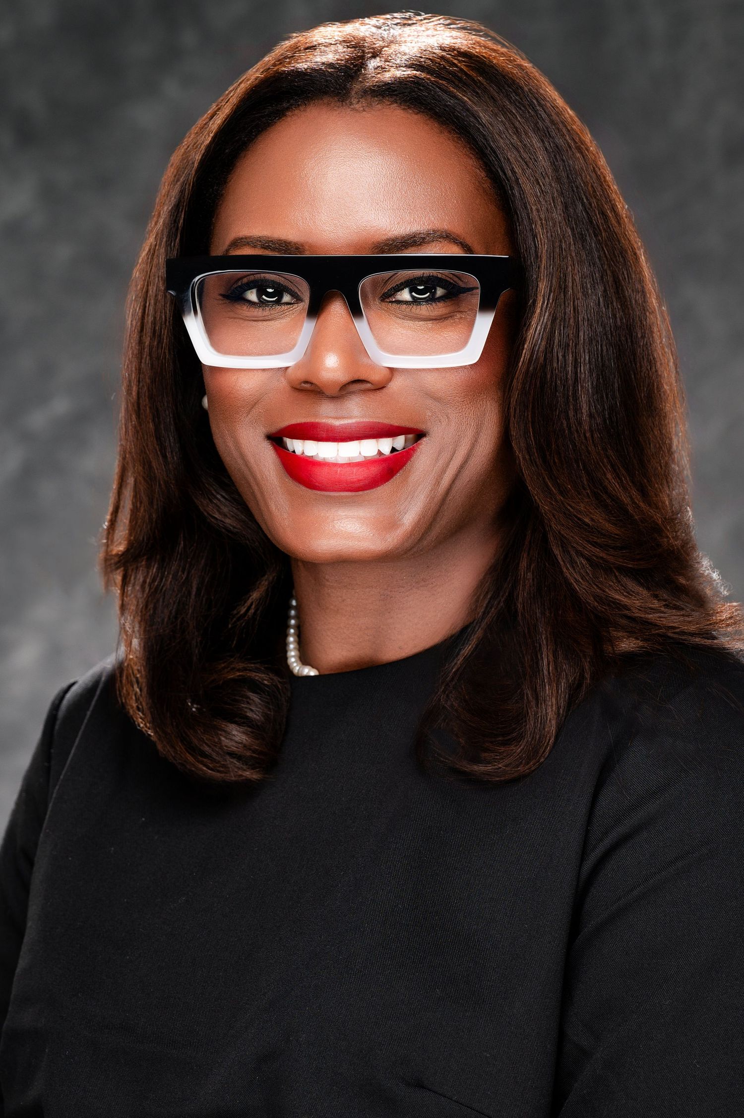Professional headshot of someone wearing a black blazer, white-framed glasses and pearl necklace against a dark background.