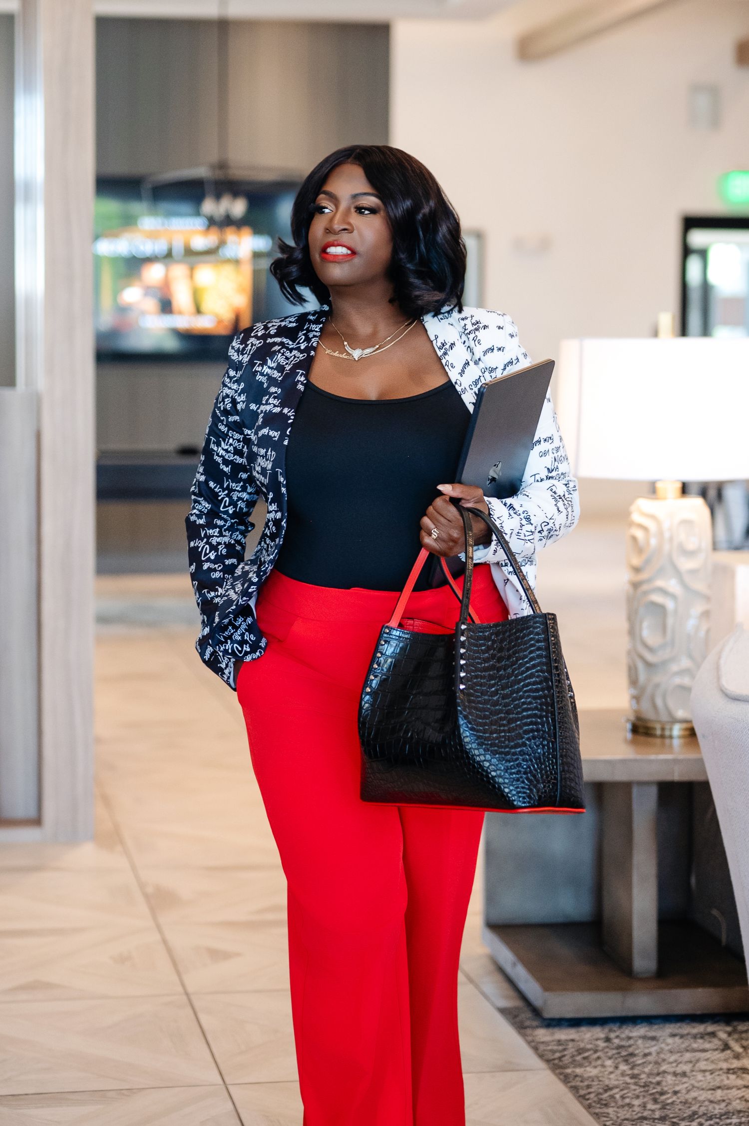 Professional Black woman in red pants, black top, patterned blazer, holding tablet and handbag in modern office lobby.