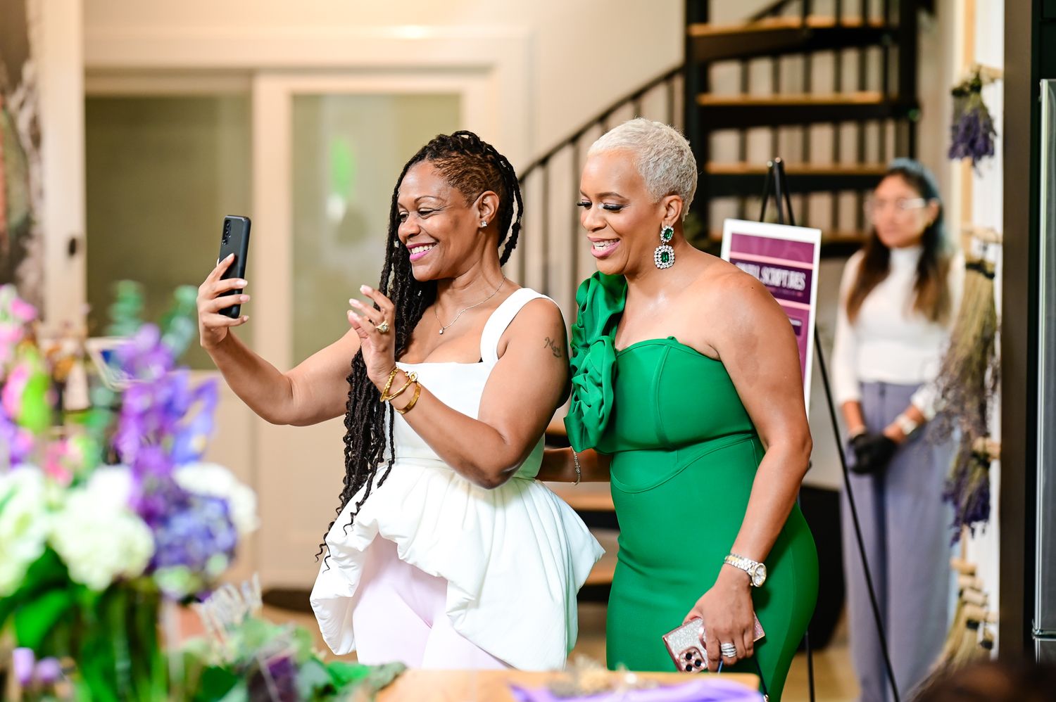 Two women smile while taking a selfie at an elegant indoor event, one in white dress, other in green outfit.