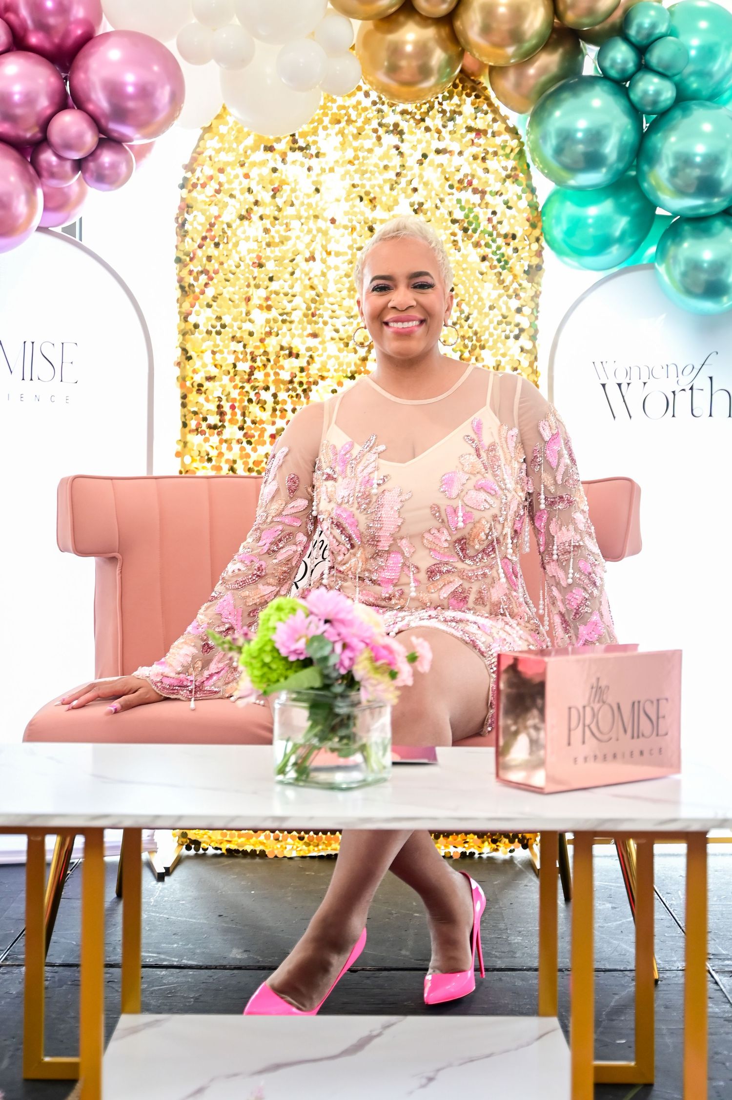A pink and gold themed celebration setup with balloon arches, decorative flowers and a pink velvet chair against a glittery backdrop.