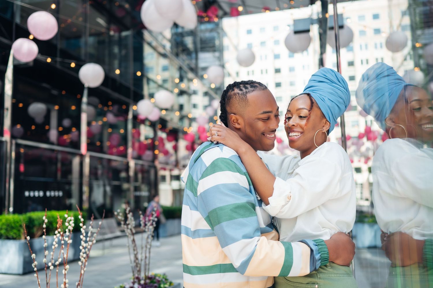 An affectionate couple embraces and smiles at an outdoor venue decorated with pink balloons and string lights.