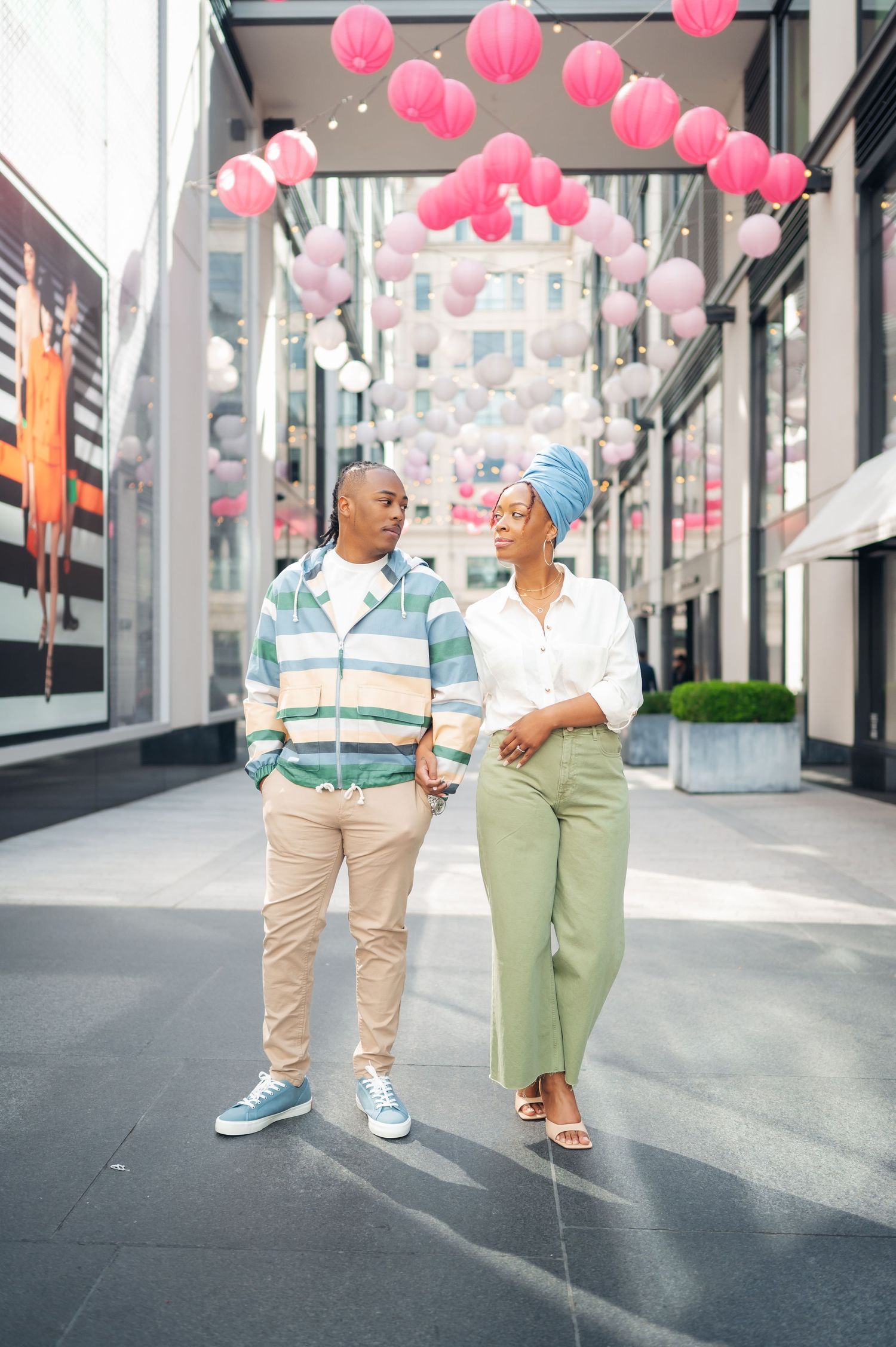 A couple walks down a modern urban walkway decorated with hanging pink lanterns overhead in a festive display.